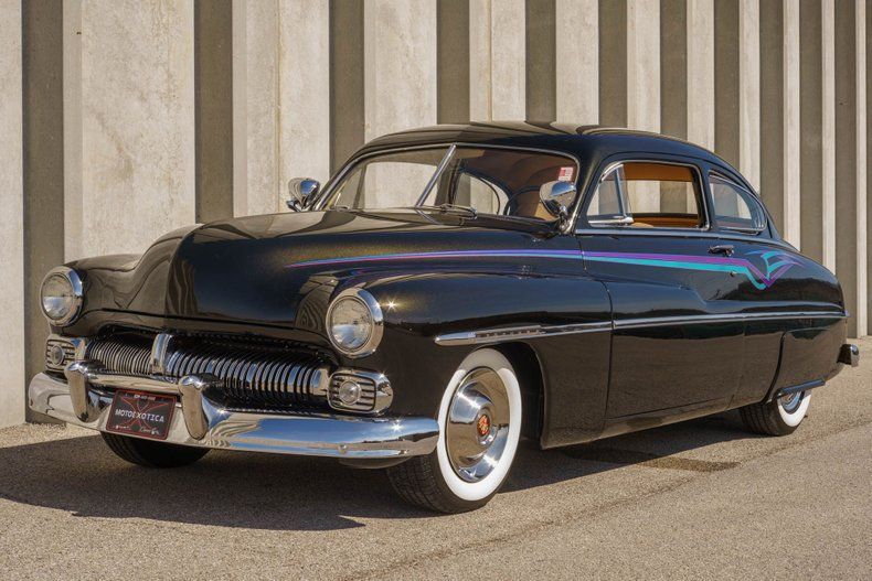 A shiny, black 1950s Mercury custom car with white-wall tires parked in front of a striped concrete wall.