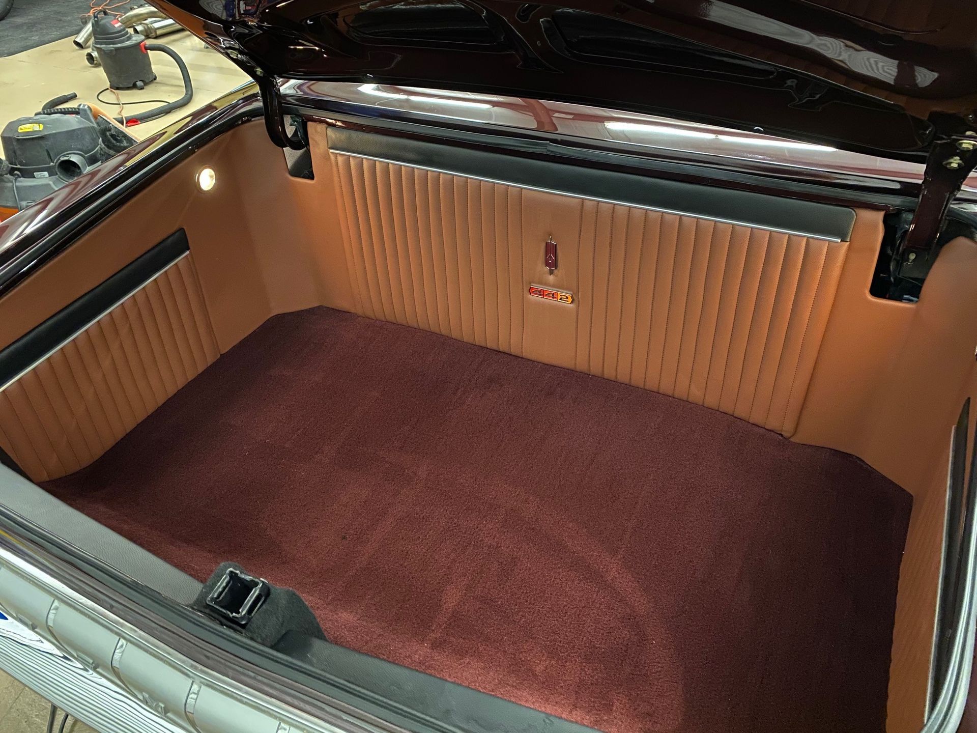A pristine car trunk with deep maroon carpeting and tan, vertically pleated custom interior side paneling.