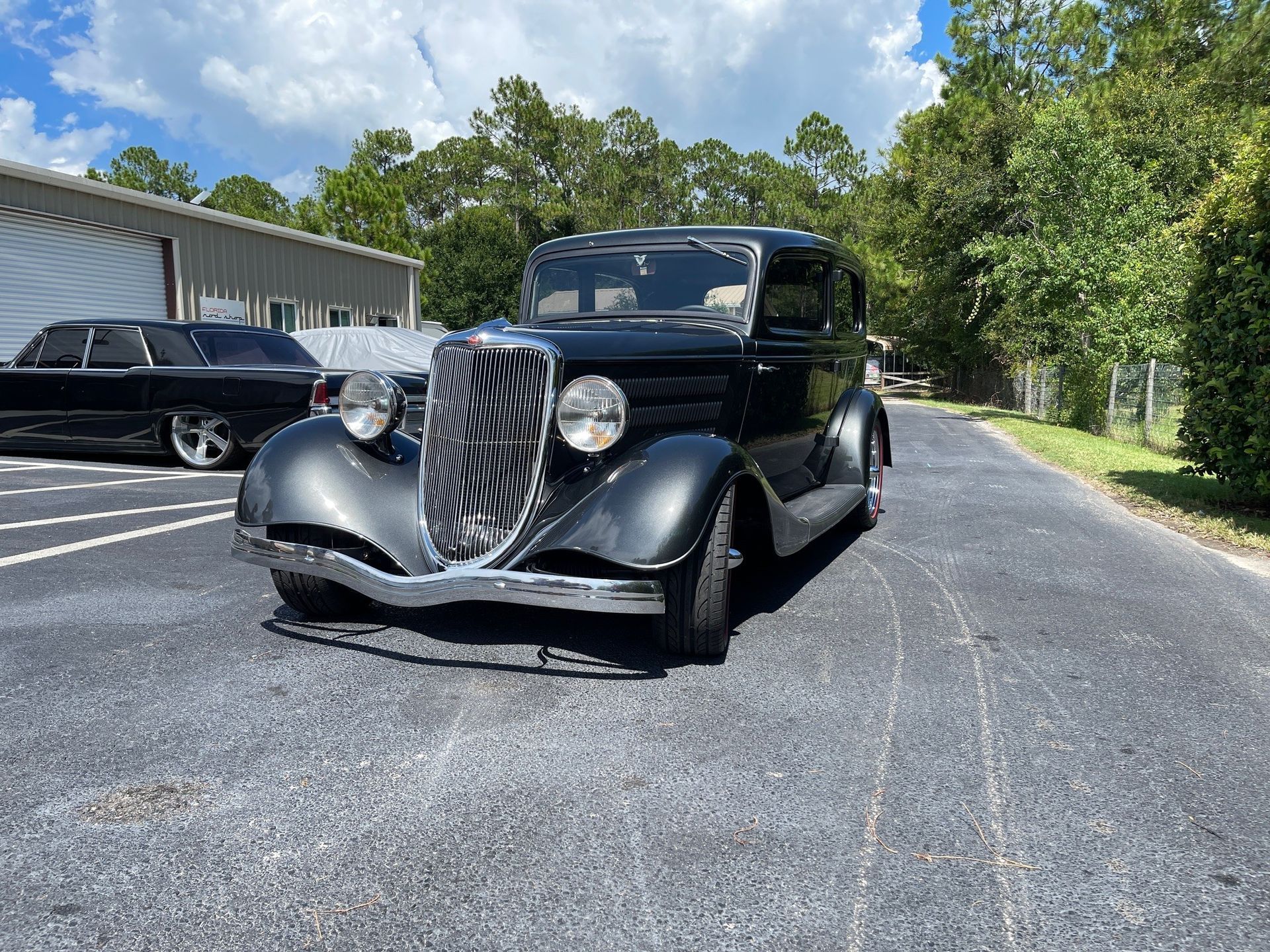 A black vintage sedan with chrome detailing parked on an asphalt lot in front of a building and trees under a blue sky.