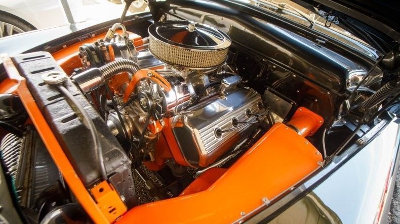 A bright orange, classic V8 car engine with polished chrome components sits in a clean engine bay.