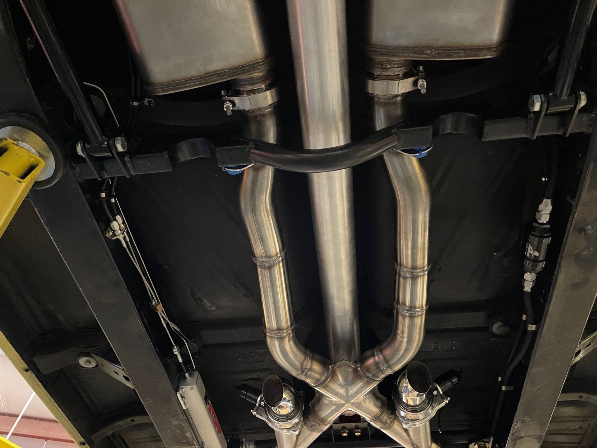 The underside of a vehicle showing an exhaust system with a metallic X-pipe connected to dual mufflers.