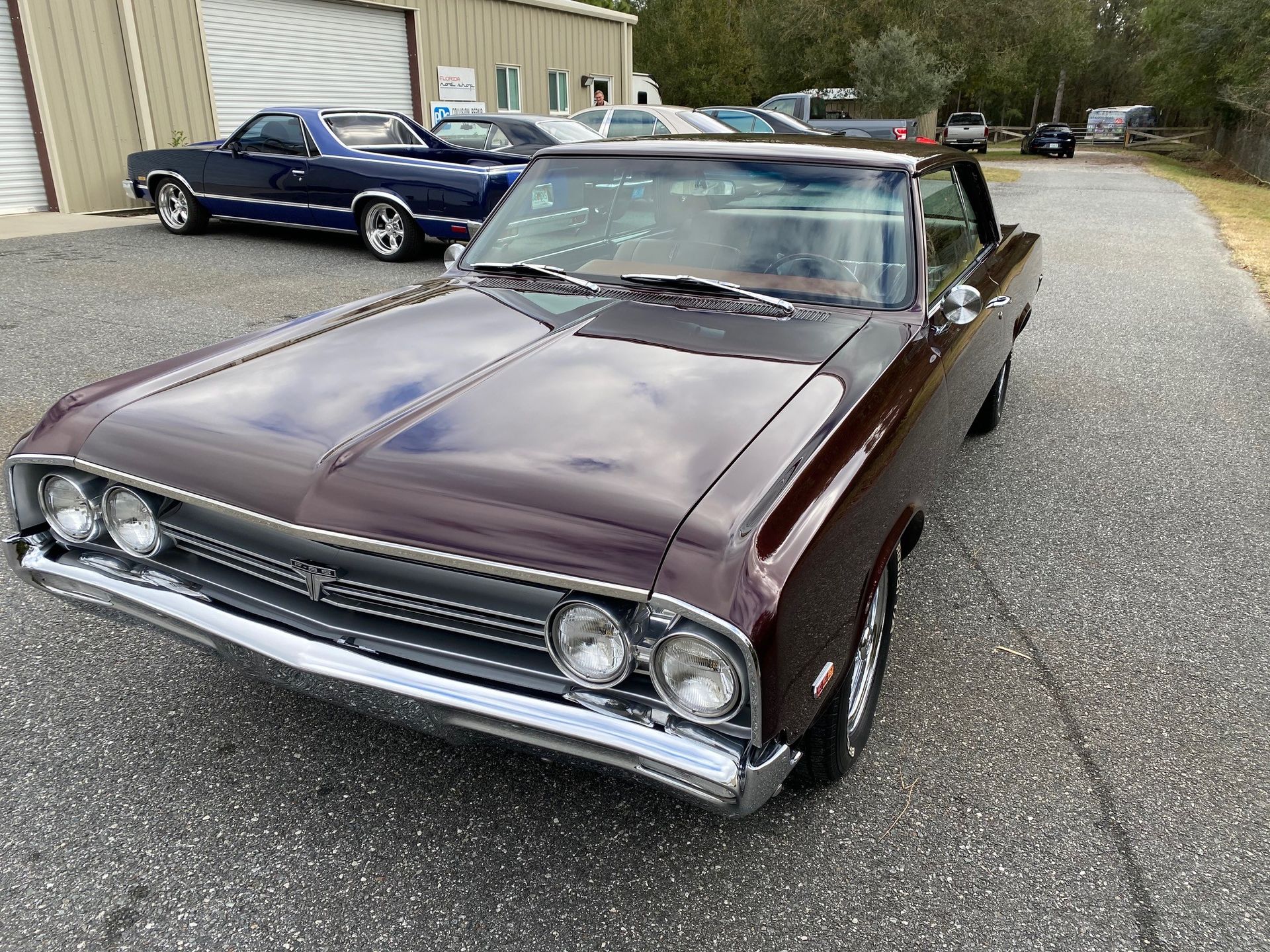 A glossy, dark maroon vintage muscle car parked on an asphalt lot in front of a warehouse.
