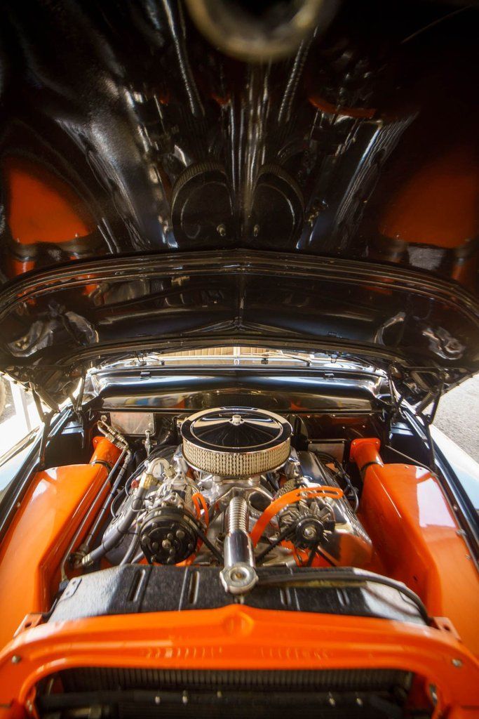 The open hood of a classic car reveals a polished chrome engine bay framed by vibrant orange fender panels.