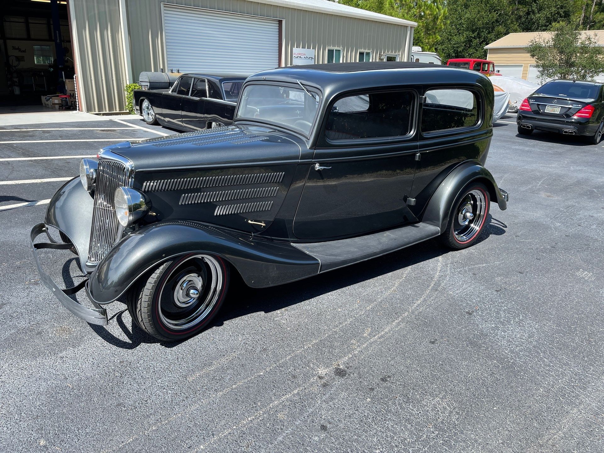 A shiny, dark gray vintage sedan parked on an asphalt lot in front of a light-colored building.