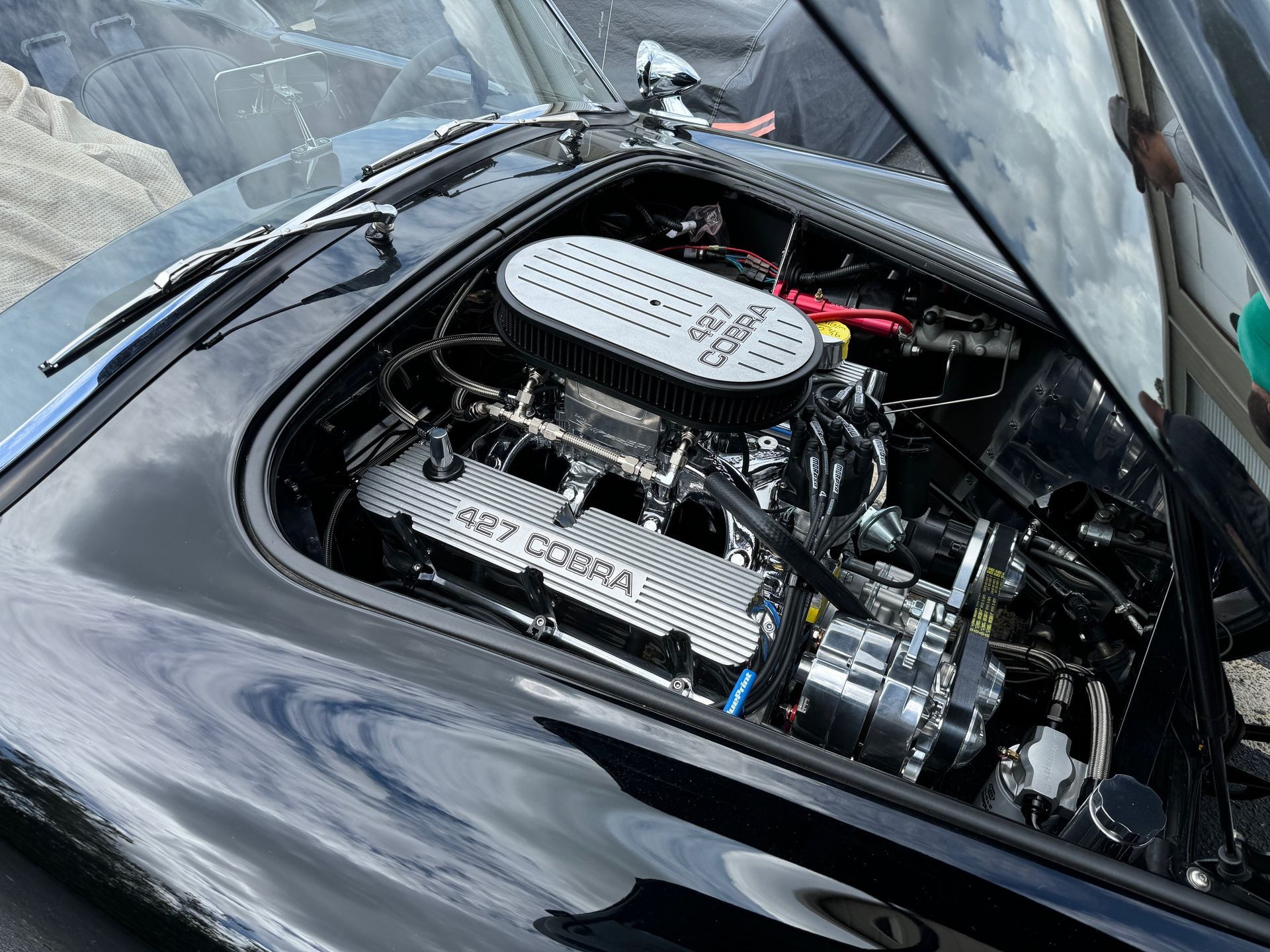 A high-angle view of a polished black car's open hood, revealing a chrome-detailed 427 Cobra engine with a finned air cover.