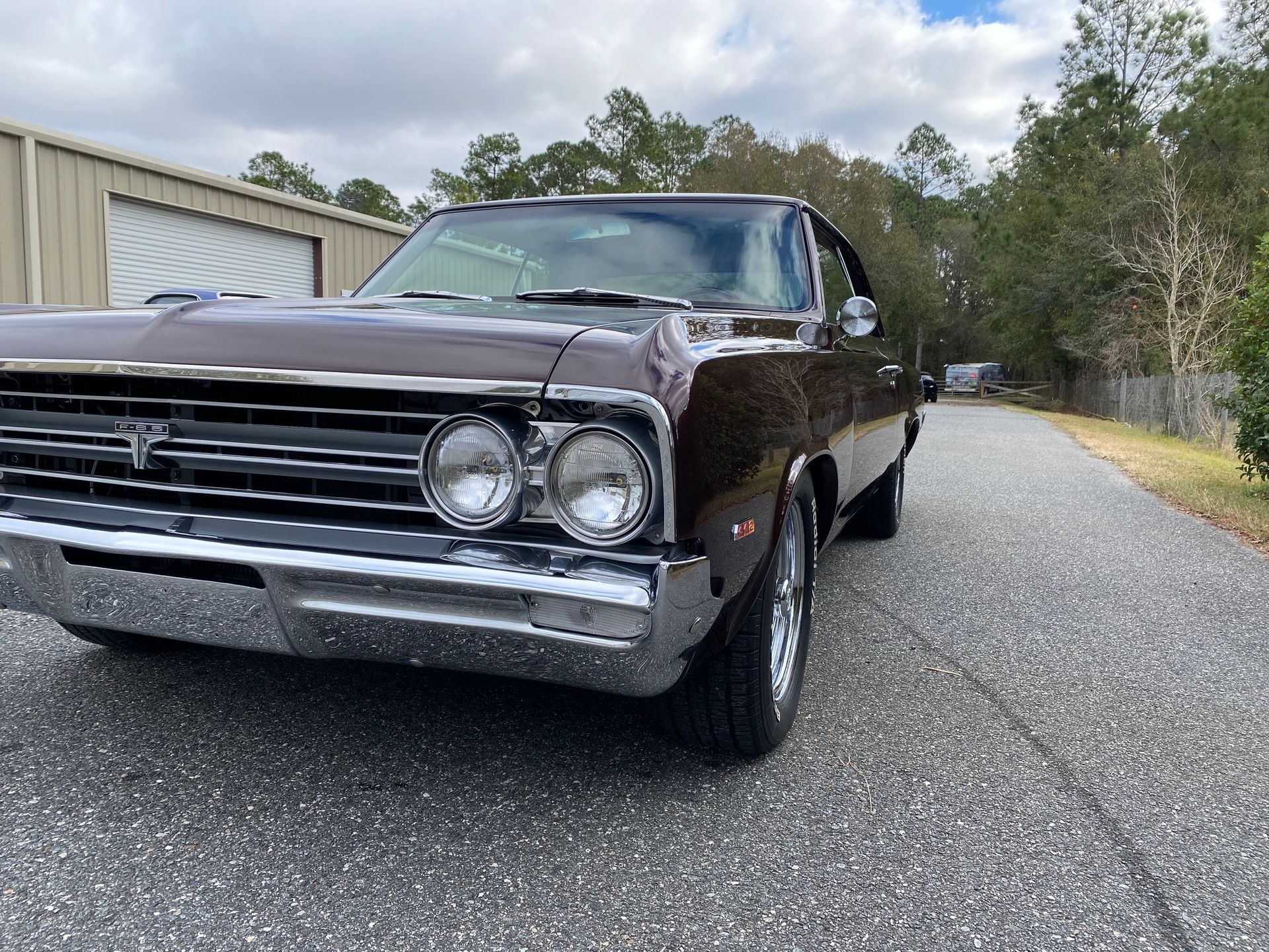 A brown vintage classic car parked on a gravel driveway in front of a metal building.