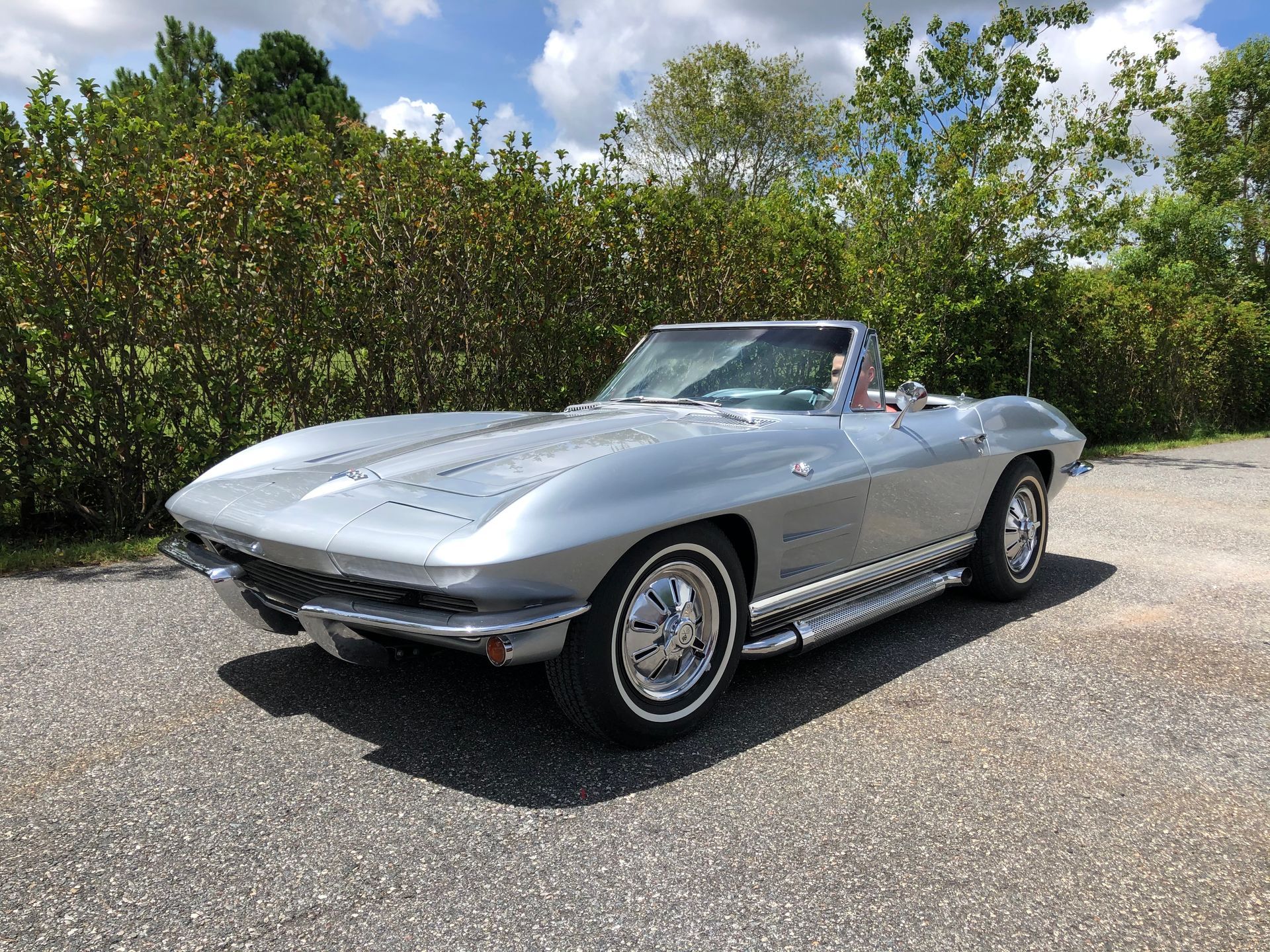 A silver classic Chevrolet Corvette convertible parked on a gravel surface in front of a green hedge.