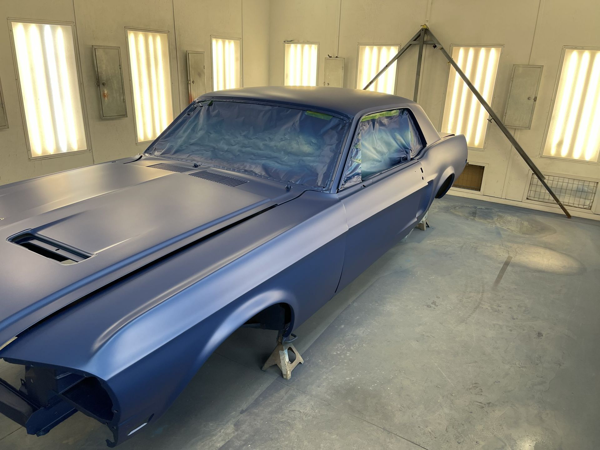 A matte navy blue classic car body on jack stands inside a brightly lit professional paint booth.