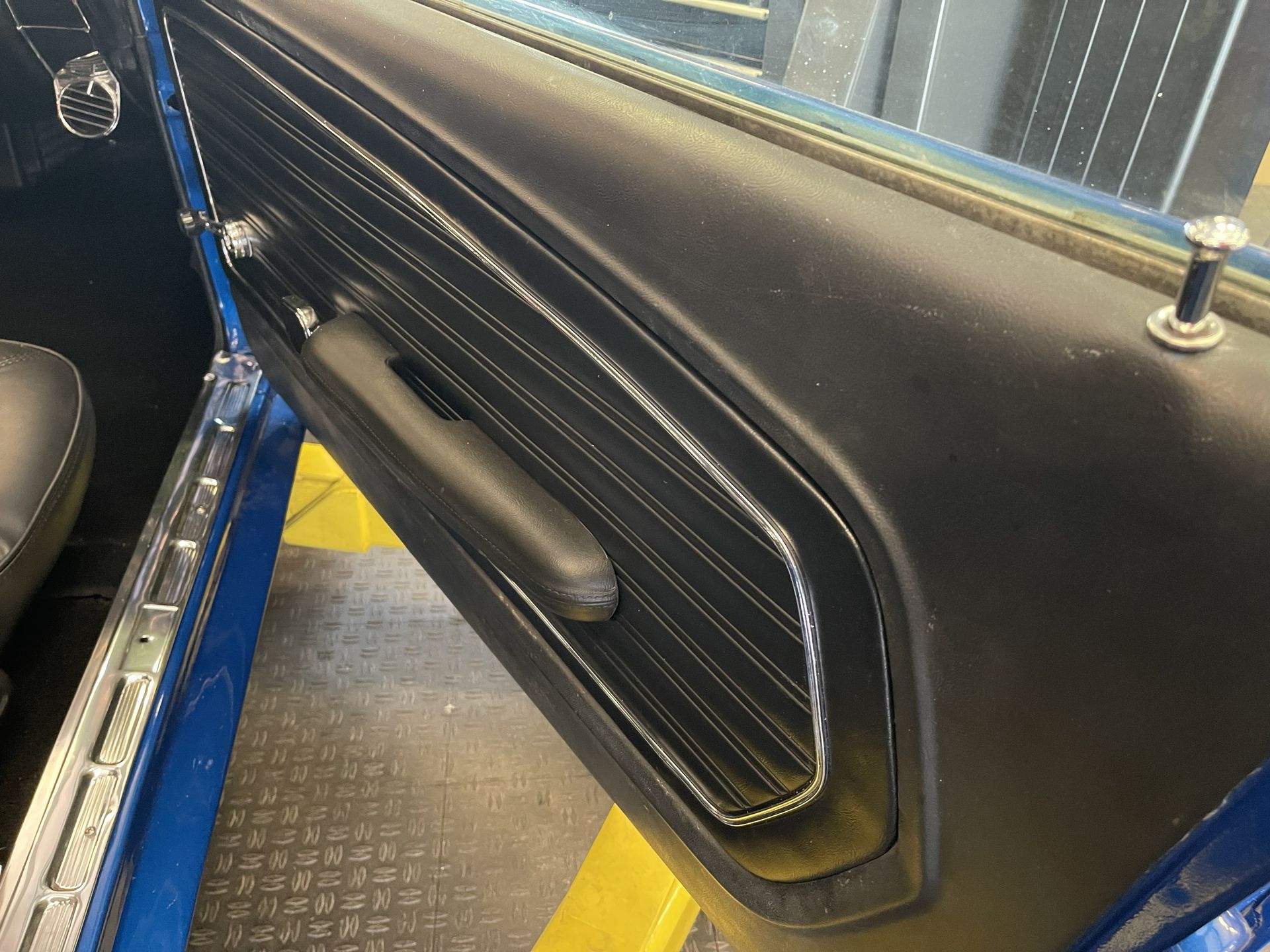 The interior of a blue car door, featuring a black ribbed panel, an armrest, and a silver lock pin.