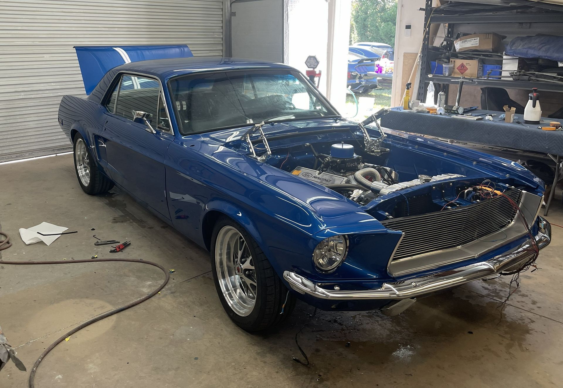 A blue vintage Ford Mustang under restoration in a garage, with the hood open, chrome wheels, and a custom grille.