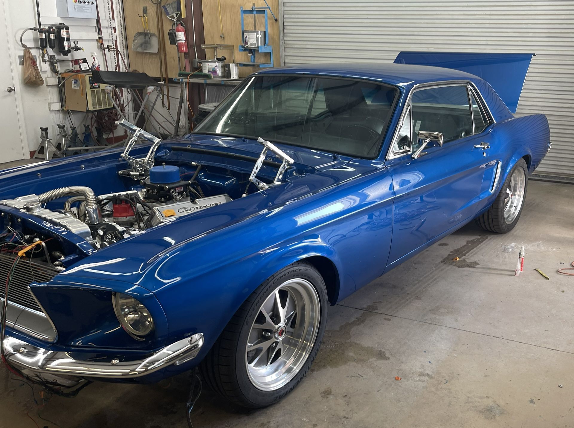 A blue vintage Ford Mustang with its hood open, parked inside a garage workshop.