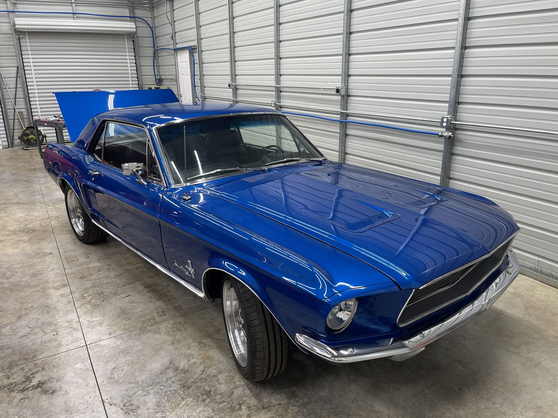 A classic bright blue Ford Mustang coupe parked inside a garage with its trunk open.