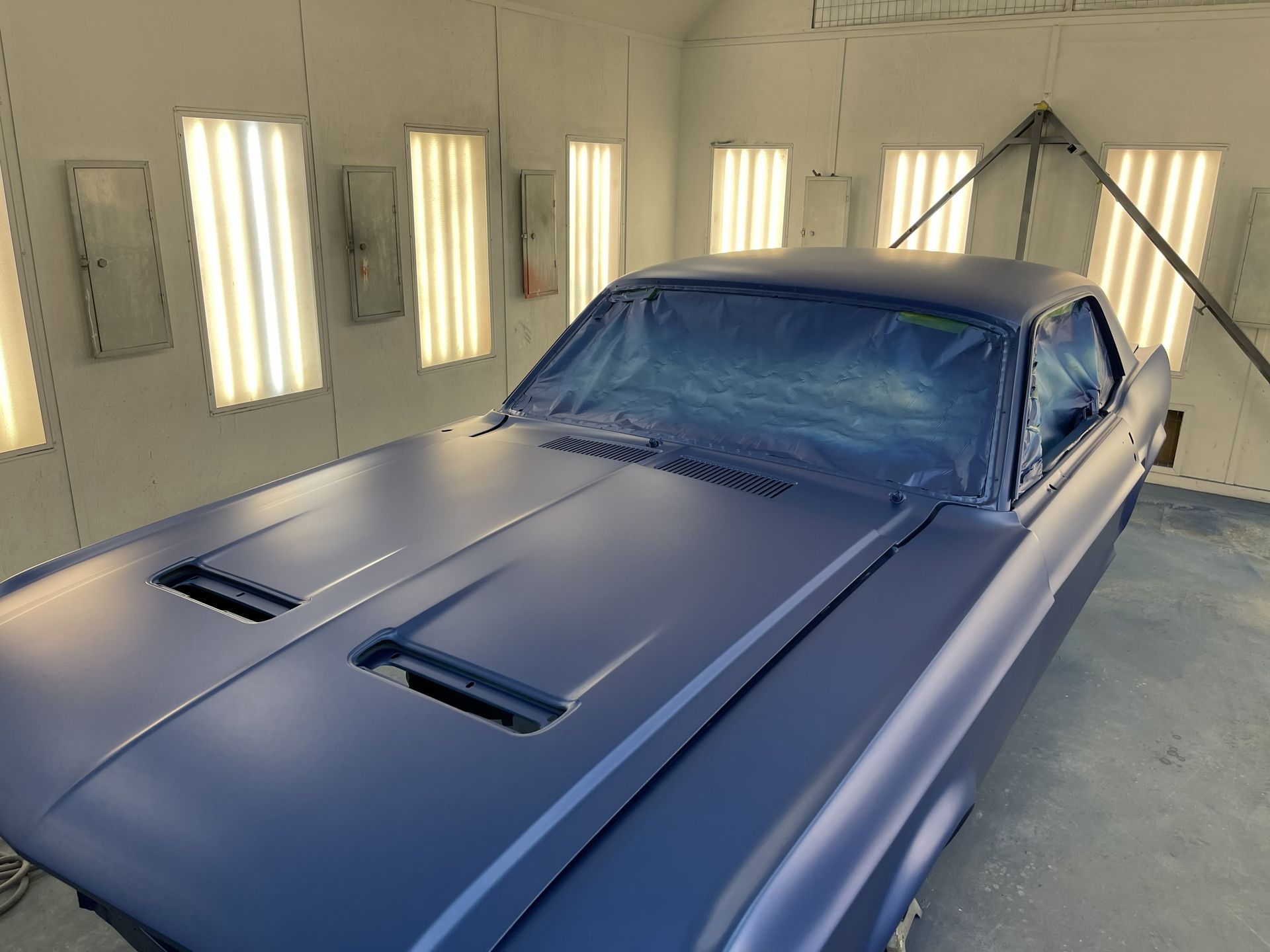 A matte blue classic car sits in a brightly lit automotive spray booth with the windows covered in blue masking plastic.