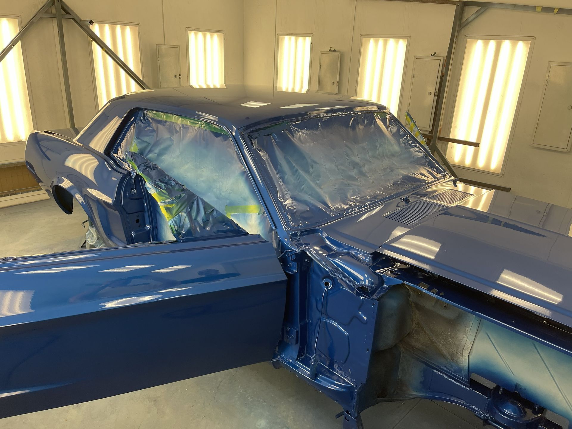 A freshly painted, glossy blue vintage car sits in an auto body shop with its door open and windows masked with plastic.