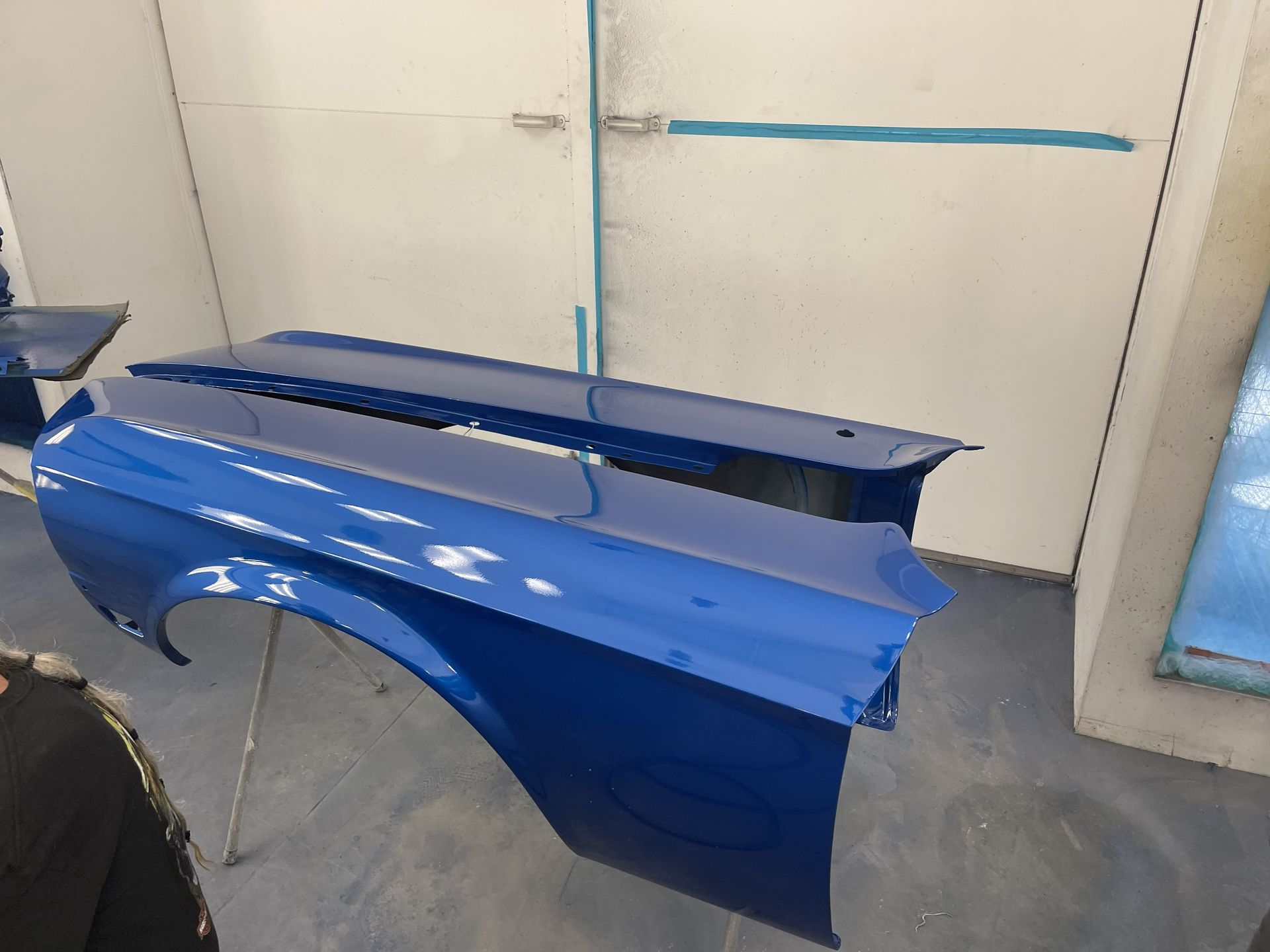 Two freshly painted royal blue car fenders drying on metal stands inside a professional paint booth.