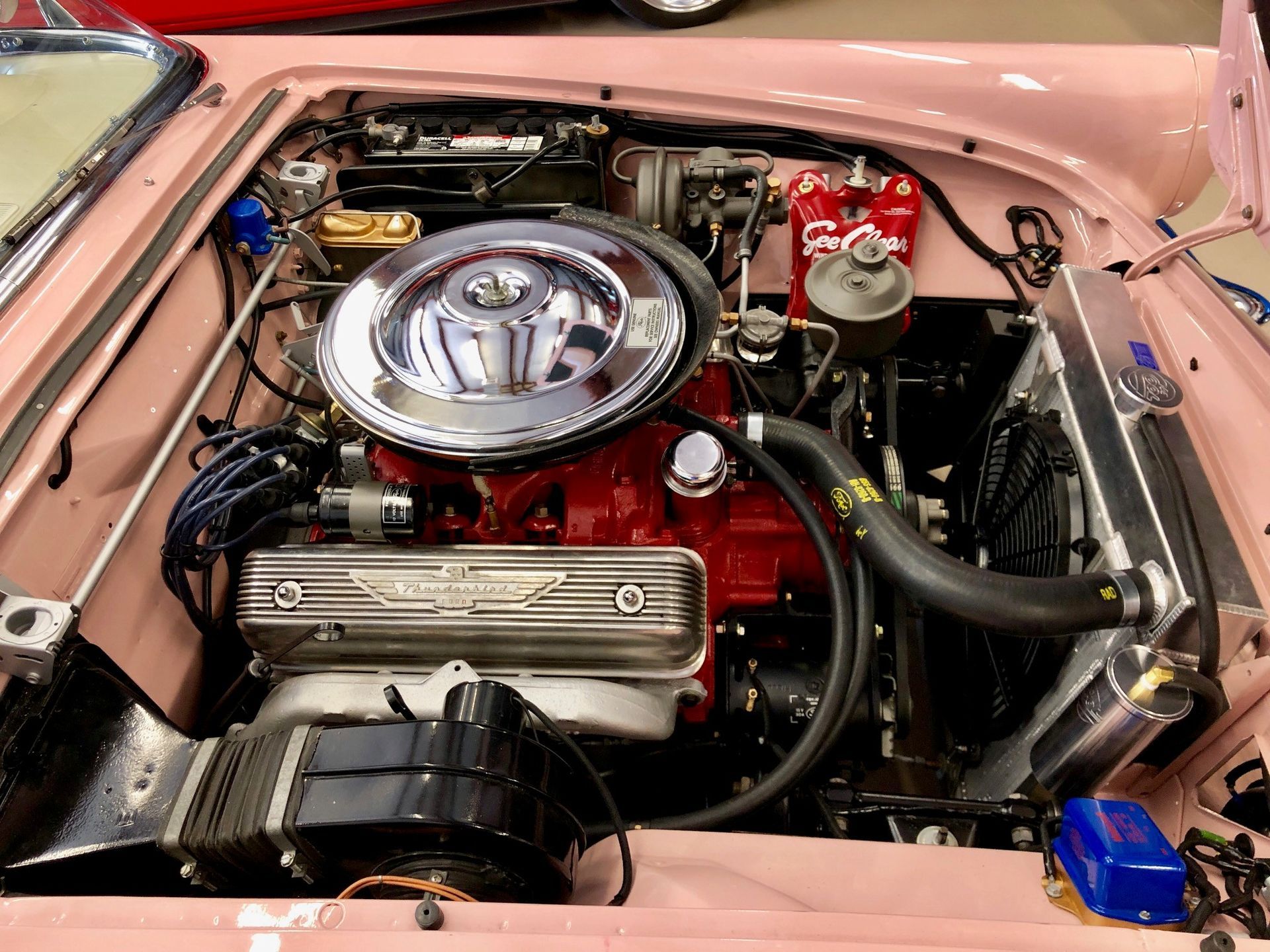 Engine bay of a vintage pink car, featuring a chrome air filter, red engine block, and metallic valve covers.