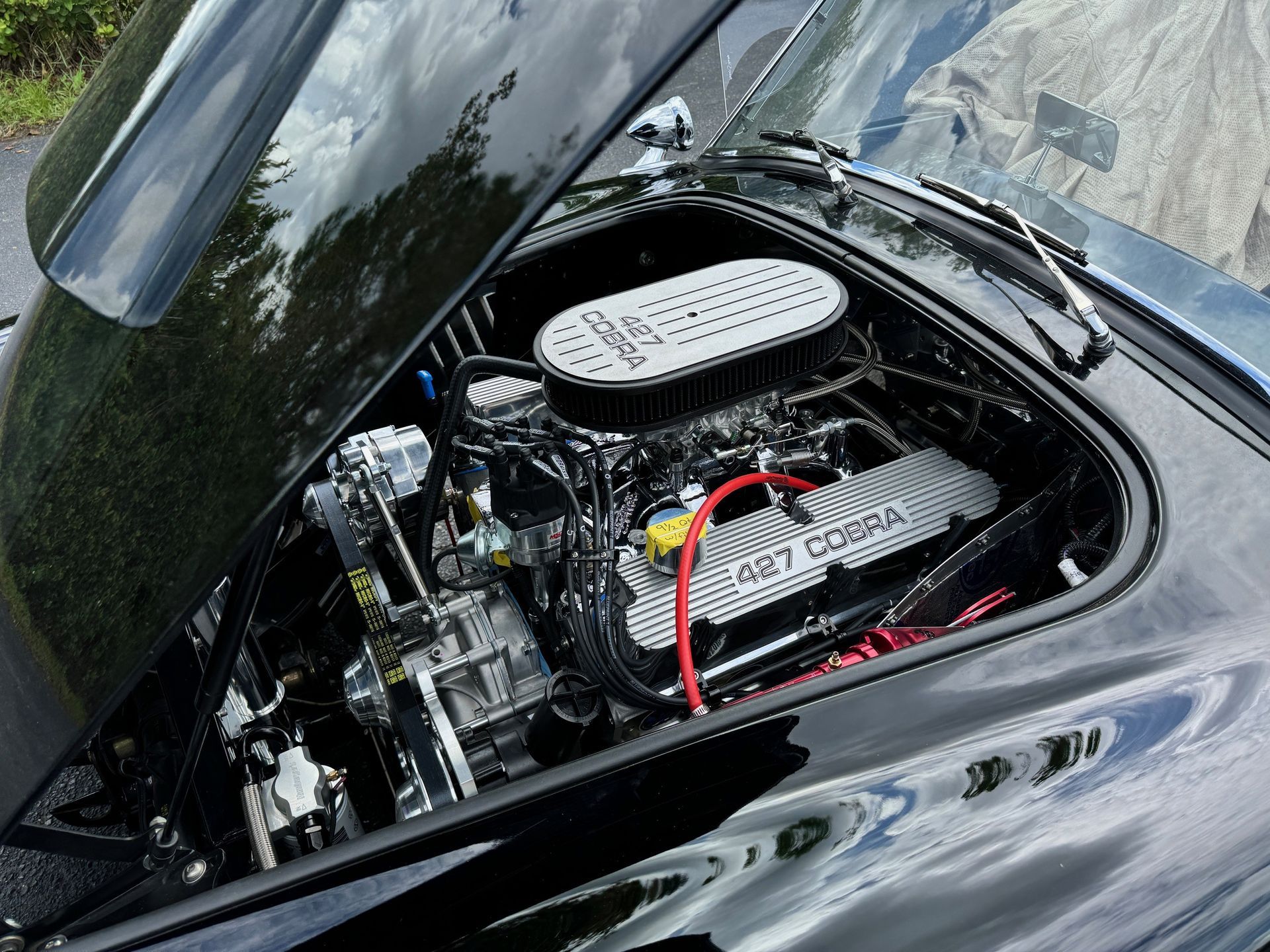 An open hood of a black car revealing a powerful 427 Cobra engine with a chrome air filter and finned valve covers.