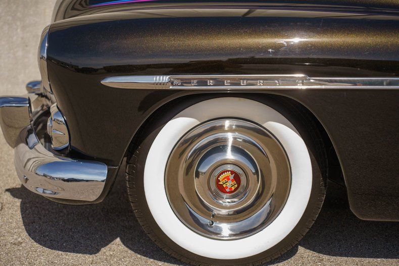 Close-up of a vintage dark brown car's front fender, chrome bumper, and a white-wall tire with a decorative hubcap.