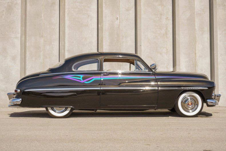A black vintage custom car with teal and purple pinstripe graphics on the side, parked against a concrete wall.
