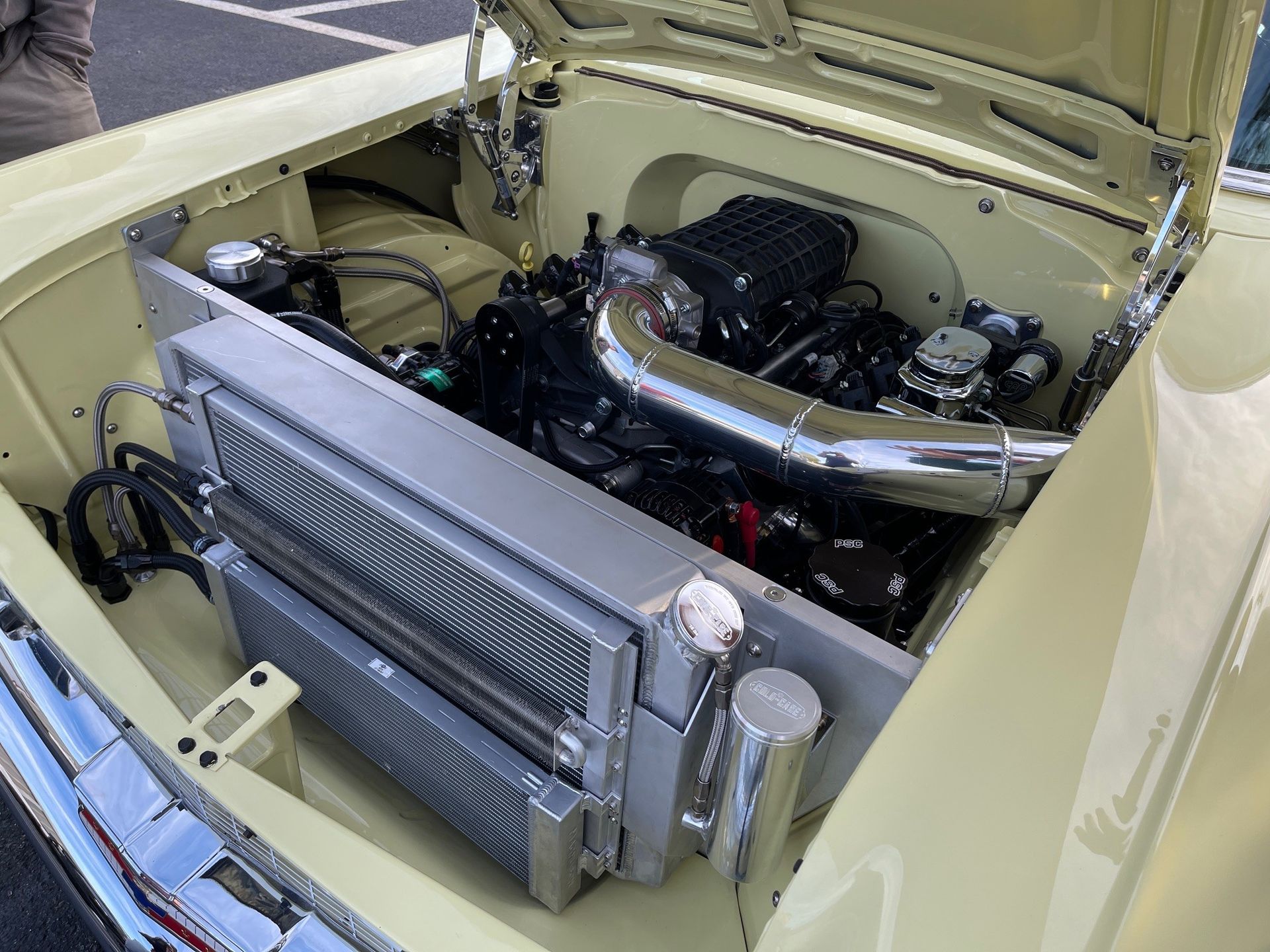 Engine bay of a vintage yellow car featuring a modern supercharged engine and a large aluminum radiator assembly.