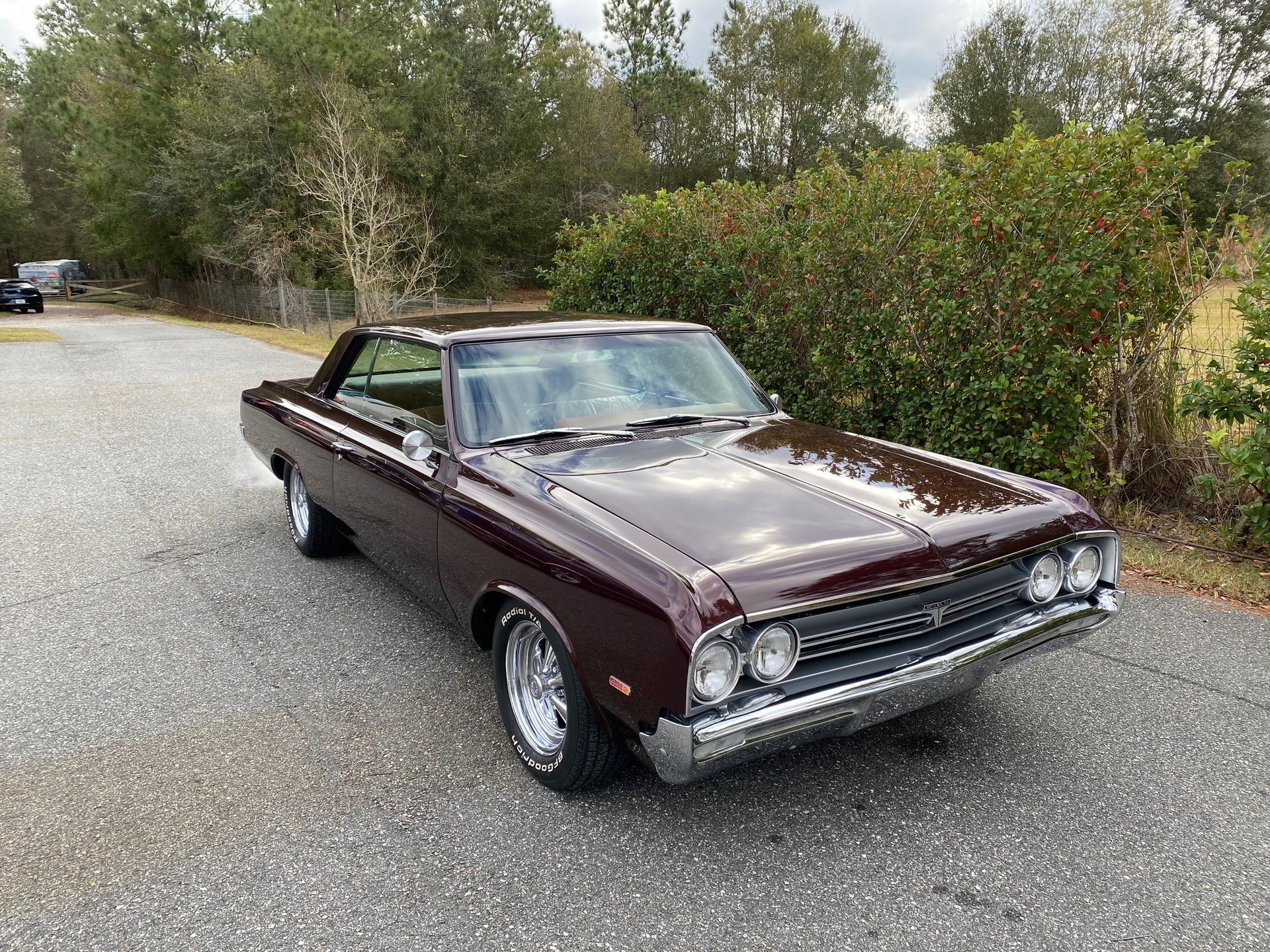 A dark maroon vintage muscle car parked on a gravel driveway surrounded by trees.