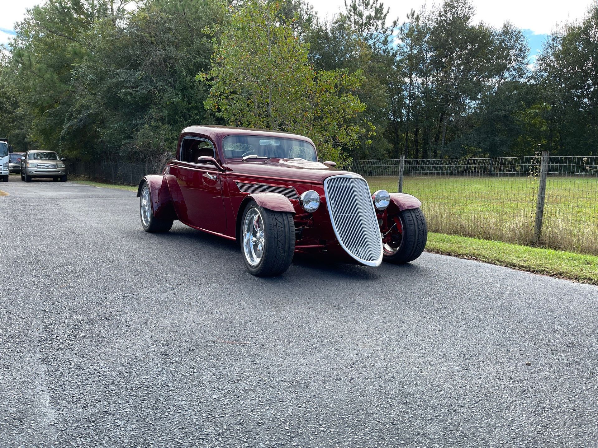 A custom maroon hot rod with a prominent chrome grille, parked on an asphalt road near trees and a field.