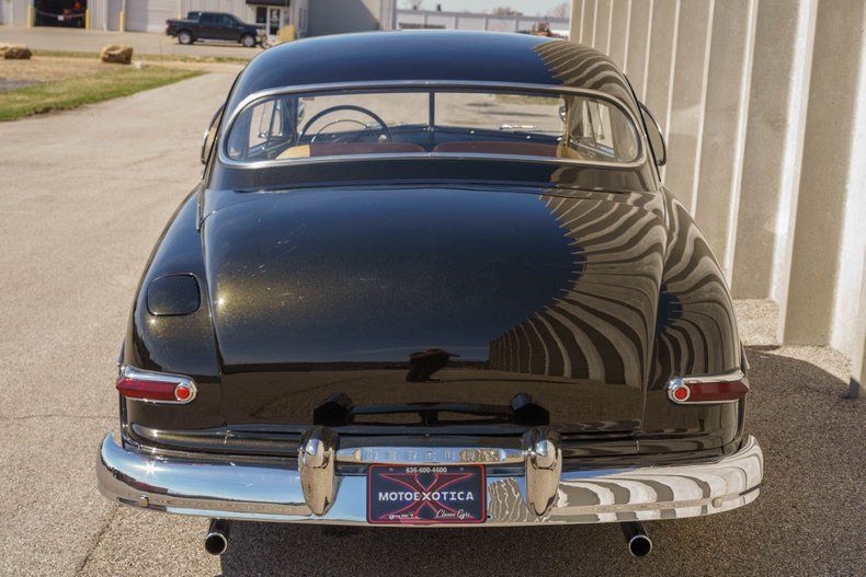The rear view of a black, vintage coupe parked on a concrete lot on a sunny day.