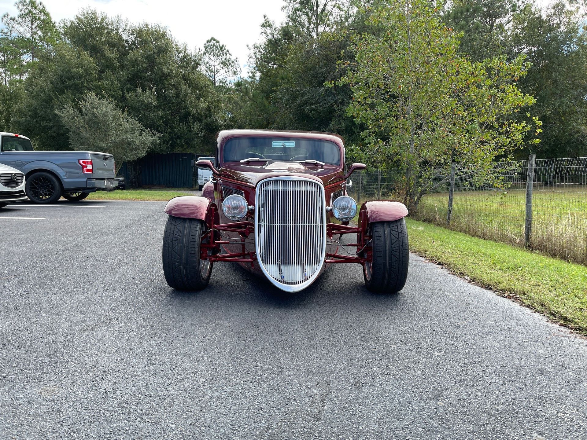 A red custom hot rod with a tall, narrow chrome grille sits on a paved lot with a gray pickup truck in the background.
