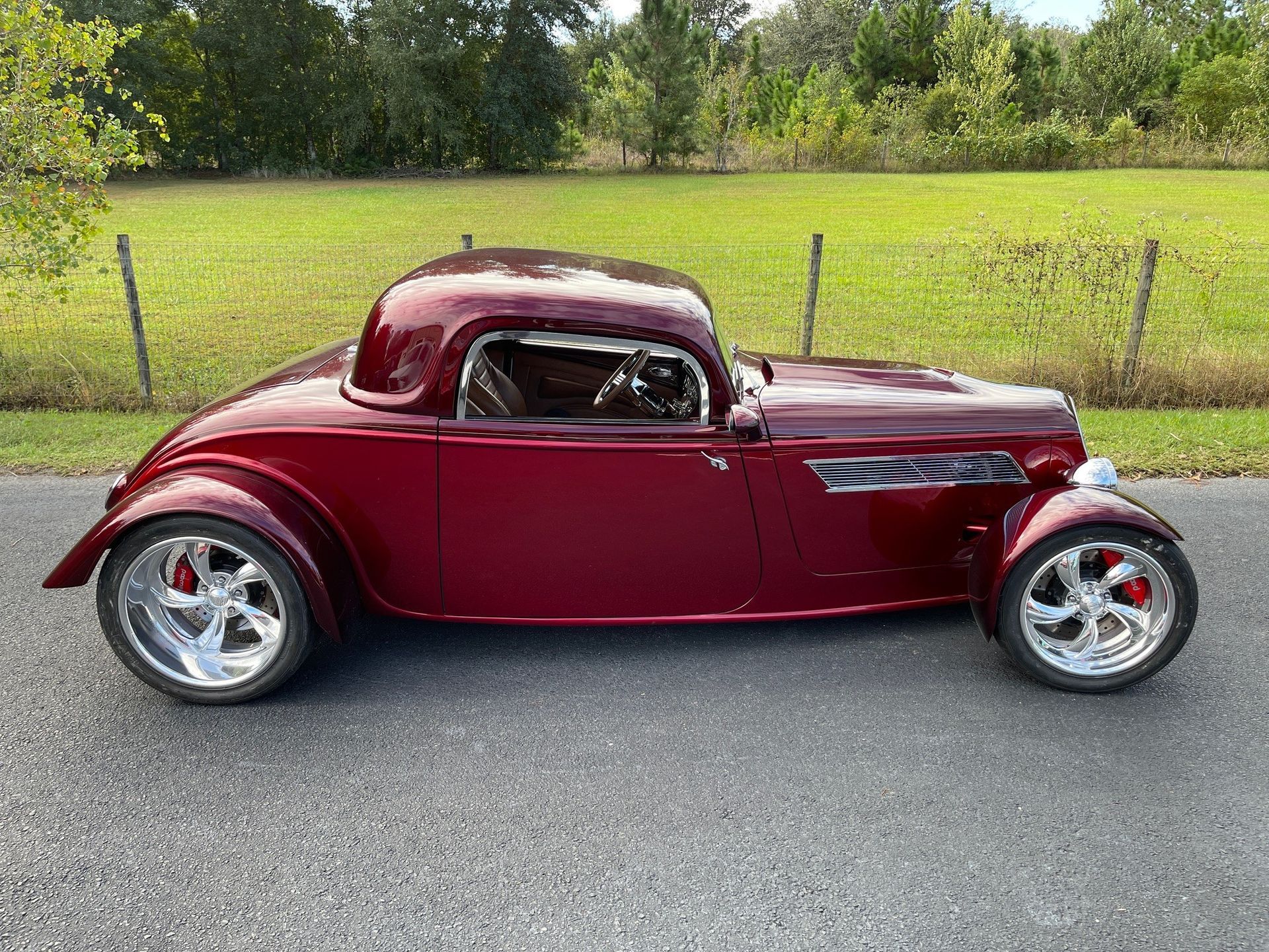A deep red vintage hot rod parked on an asphalt road in front of a green field.