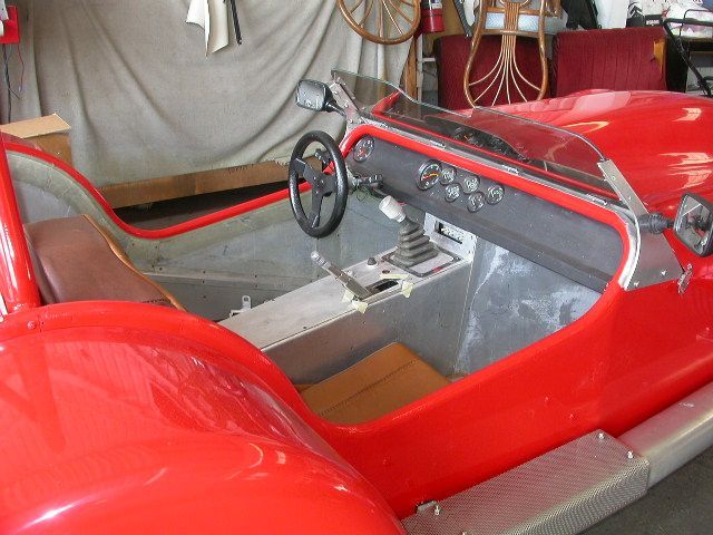 The driver's cockpit of an open-top red sports car, featuring a black steering wheel, gear shifter, and dashboard gauges.