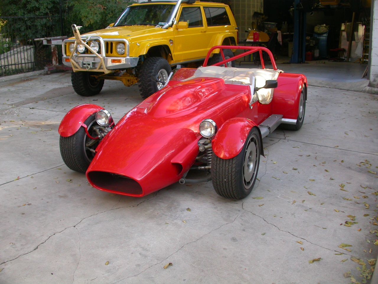 A bright red open-wheel roadster parked on a concrete driveway next to a yellow SUV.