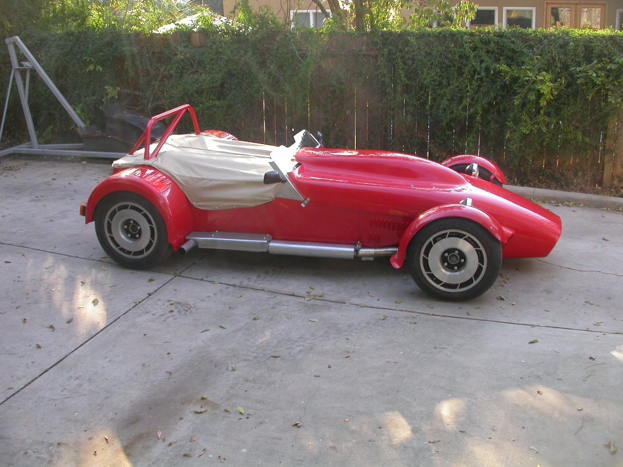 A red, vintage-style sports car with a tan tonneau cover parked on a concrete driveway.