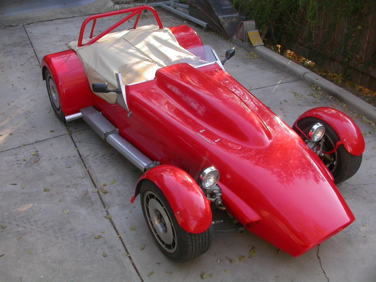 A bright red, two-seater kit car with a roll bar, tan tonneau cover, and exposed wheels on a concrete driveway.