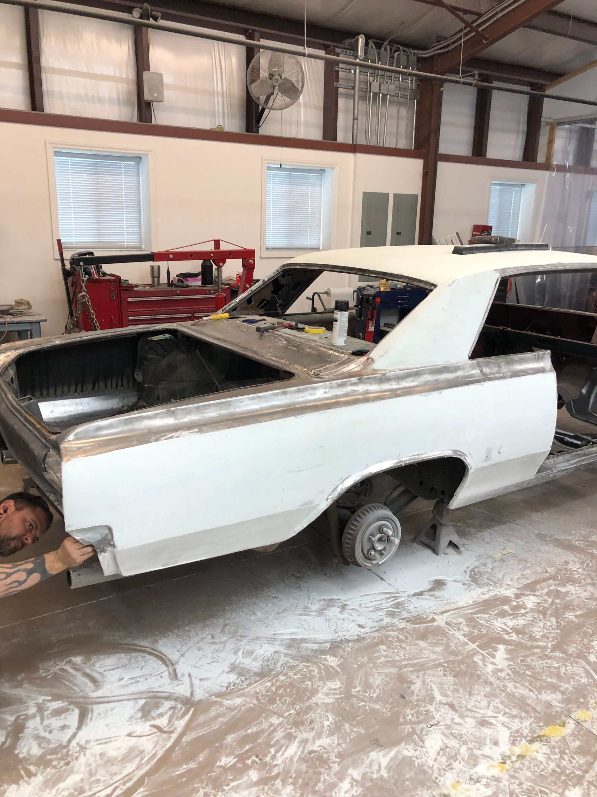 A worker sands the rear quarter panel of a white, stripped-down vintage car frame in an auto body workshop.
