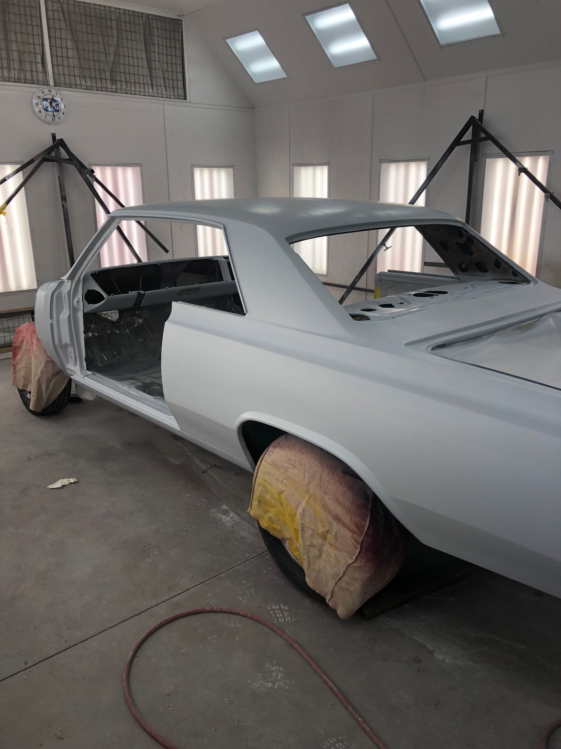 A gray car frame sits inside a brightly lit professional auto body shop, with tires covered for protection.