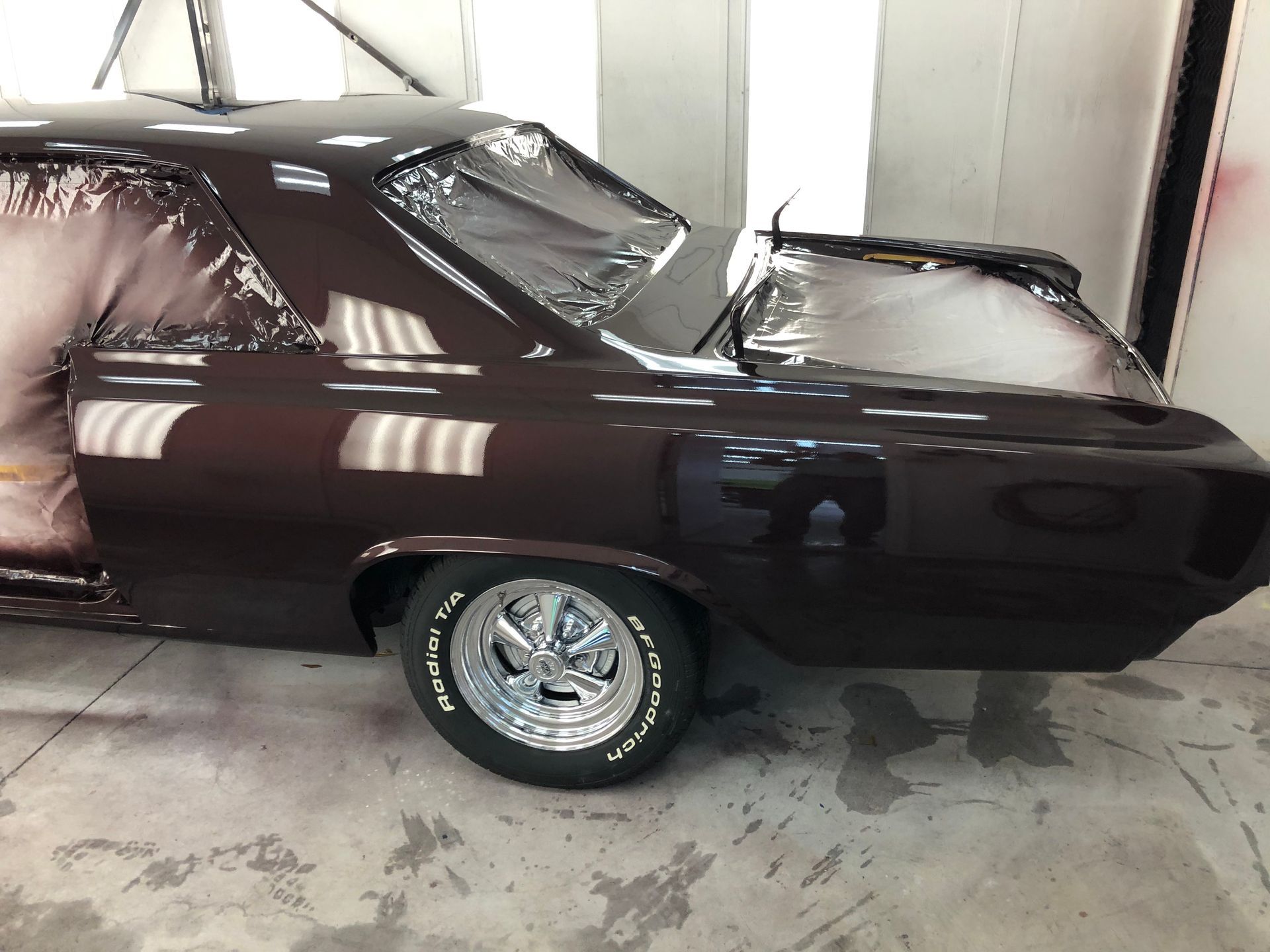 A dark brown vintage car being painted inside an auto shop, with the windows masked off and chrome wheels visible.