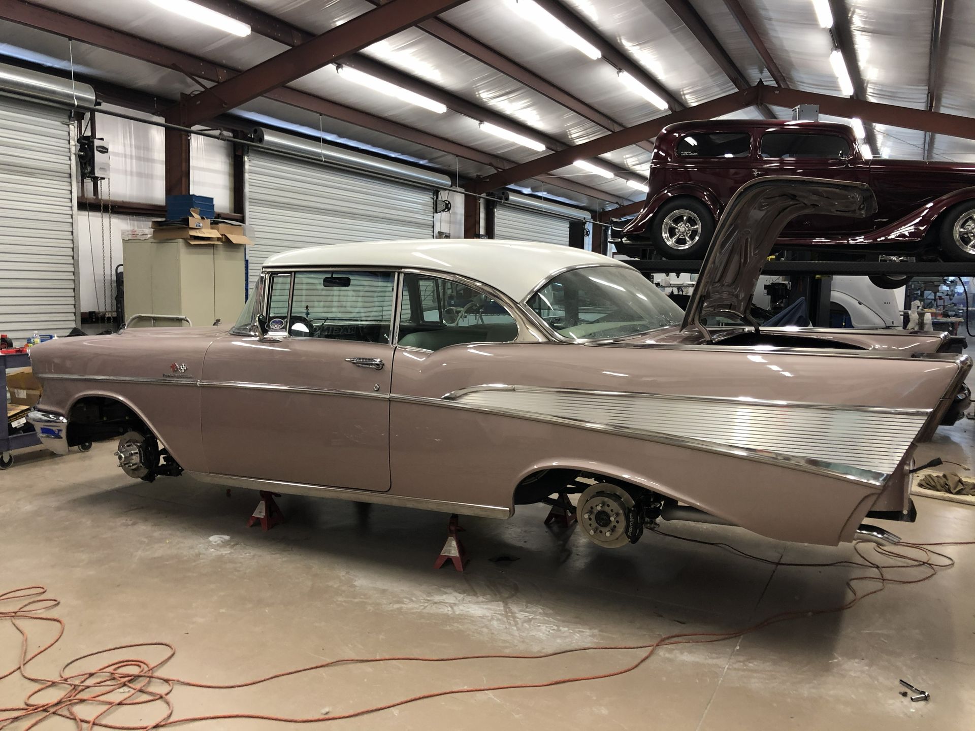 A dusty pink and white 1957 Chevrolet Bel Air rests on jack stands inside a spacious auto workshop.