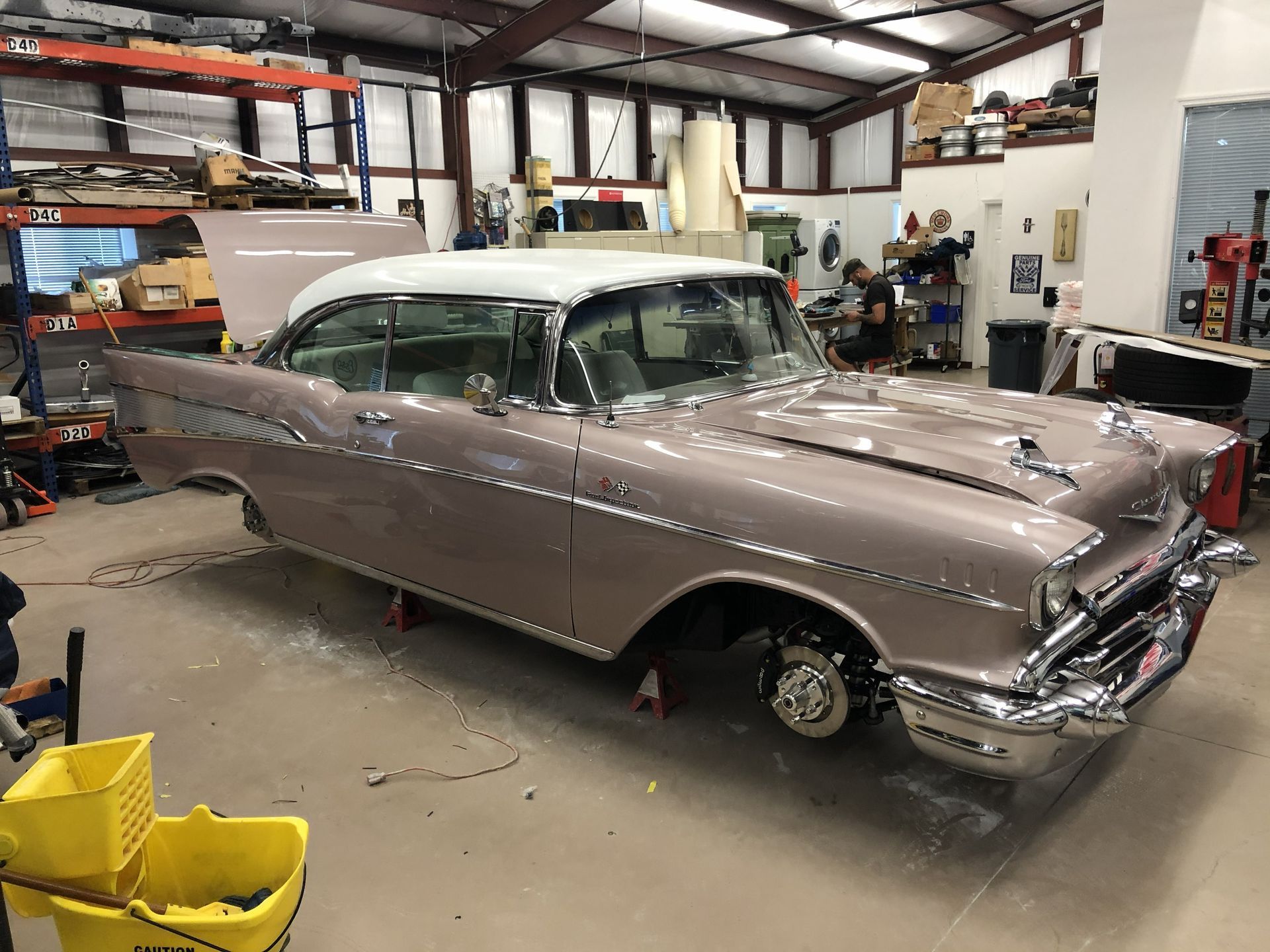 A dusty mauve and white 1957 Chevrolet Bel Air parked on jack stands inside a spacious, industrial auto repair workshop.