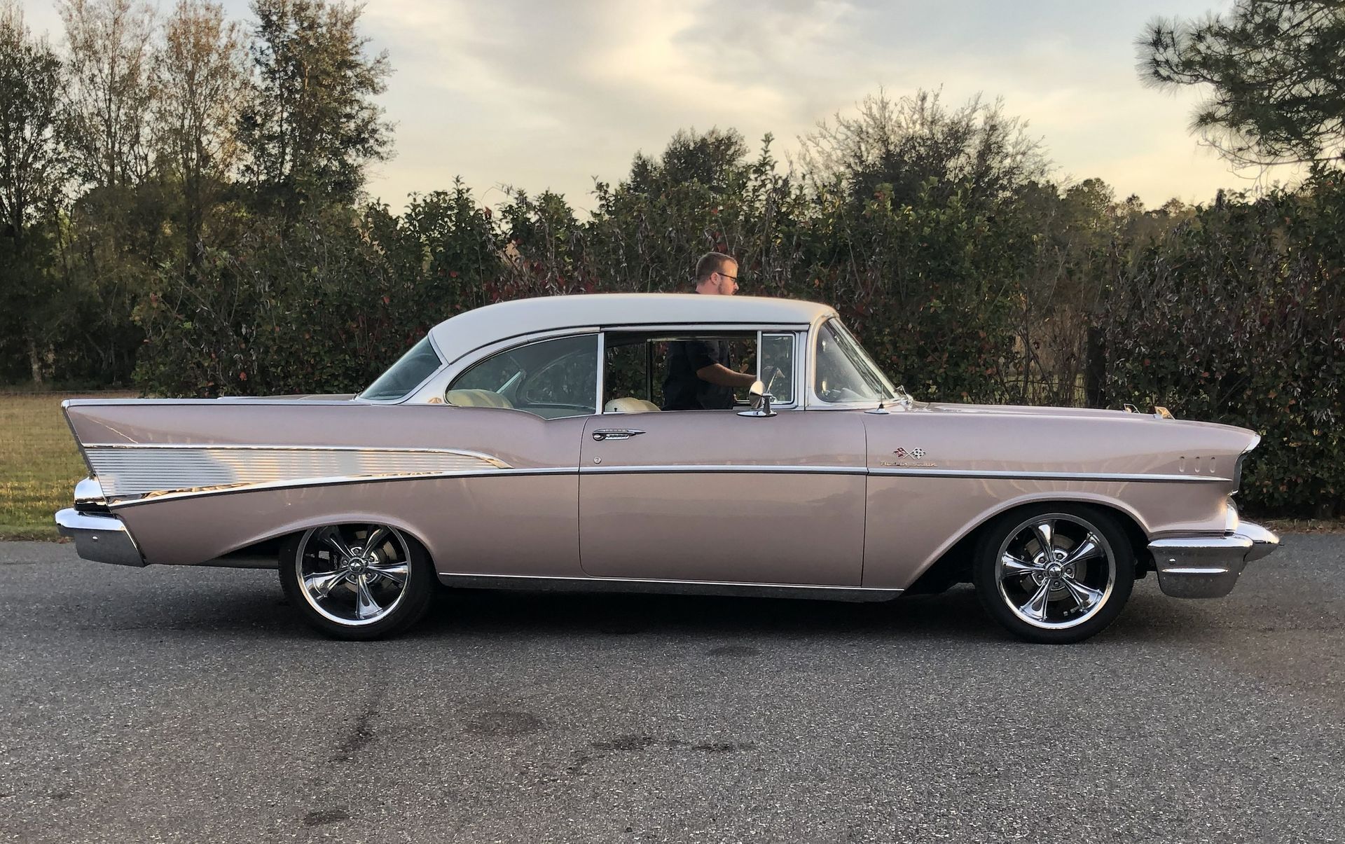 A side view of a vintage, light pink Chevrolet Bel Air parked on asphalt with a person sitting in the driver's seat.