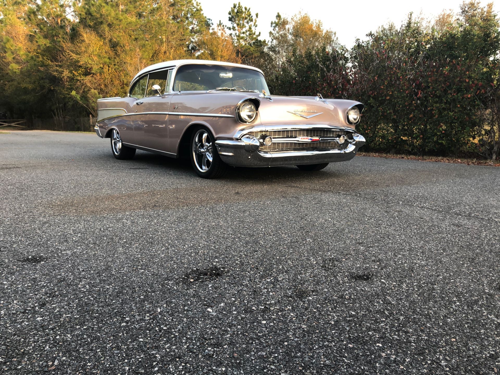 A light purple and white classic 1957 Chevrolet Bel Air parked on an asphalt driveway with trees in the background.