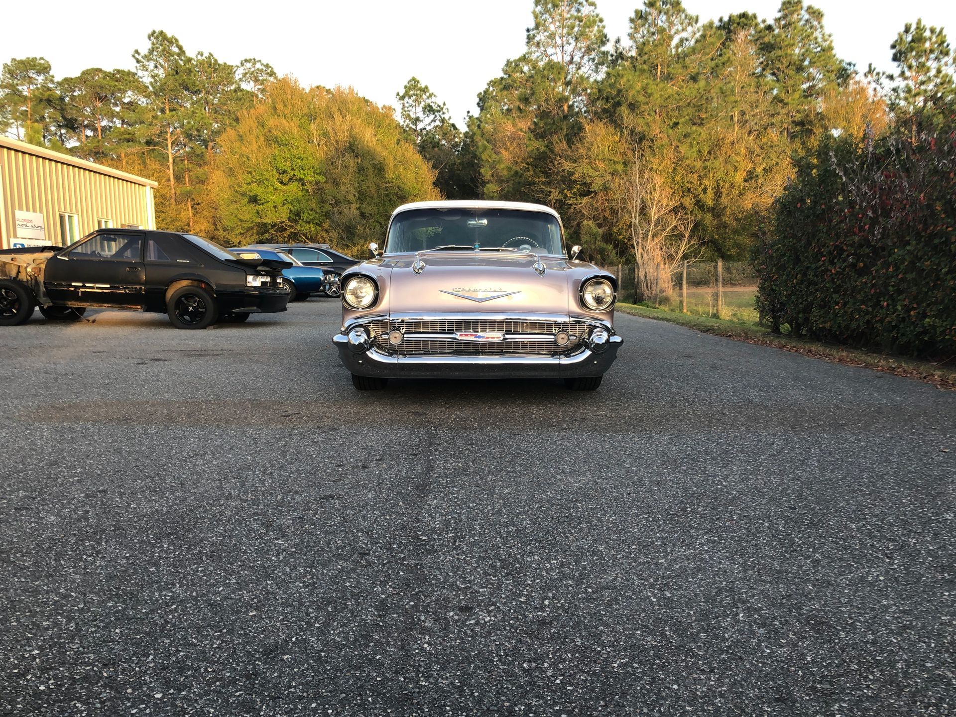 A vintage, light-colored Chevrolet Bel Air parked on a paved lot at sunset, with other cars visible in the background.