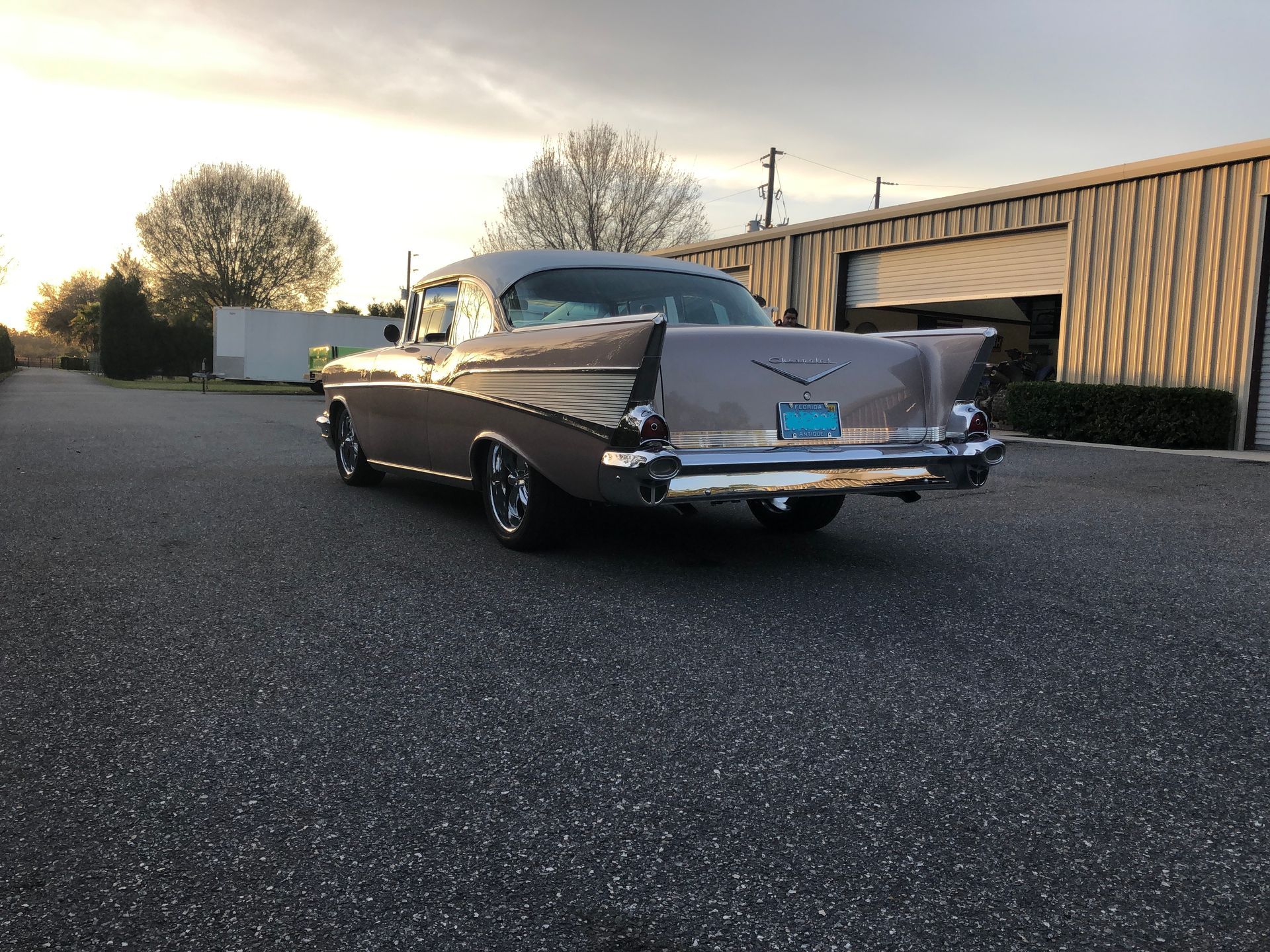 A beige 1957 Chevrolet Bel Air parked on an asphalt lot in front of a tan metal building at sunset.