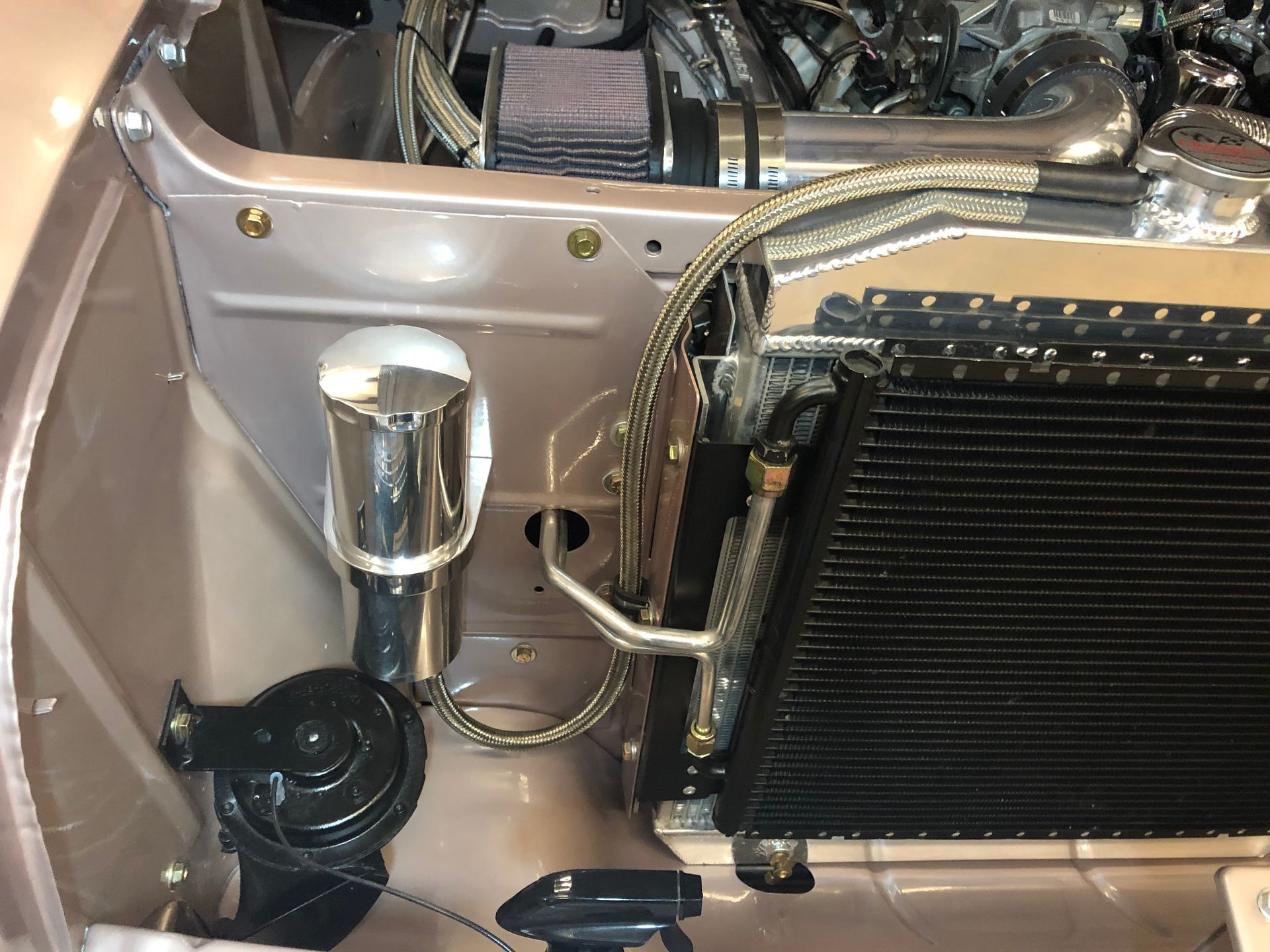 A close-up of a car engine bay showing a radiator, polished overflow tank, braided lines, and a black horn.