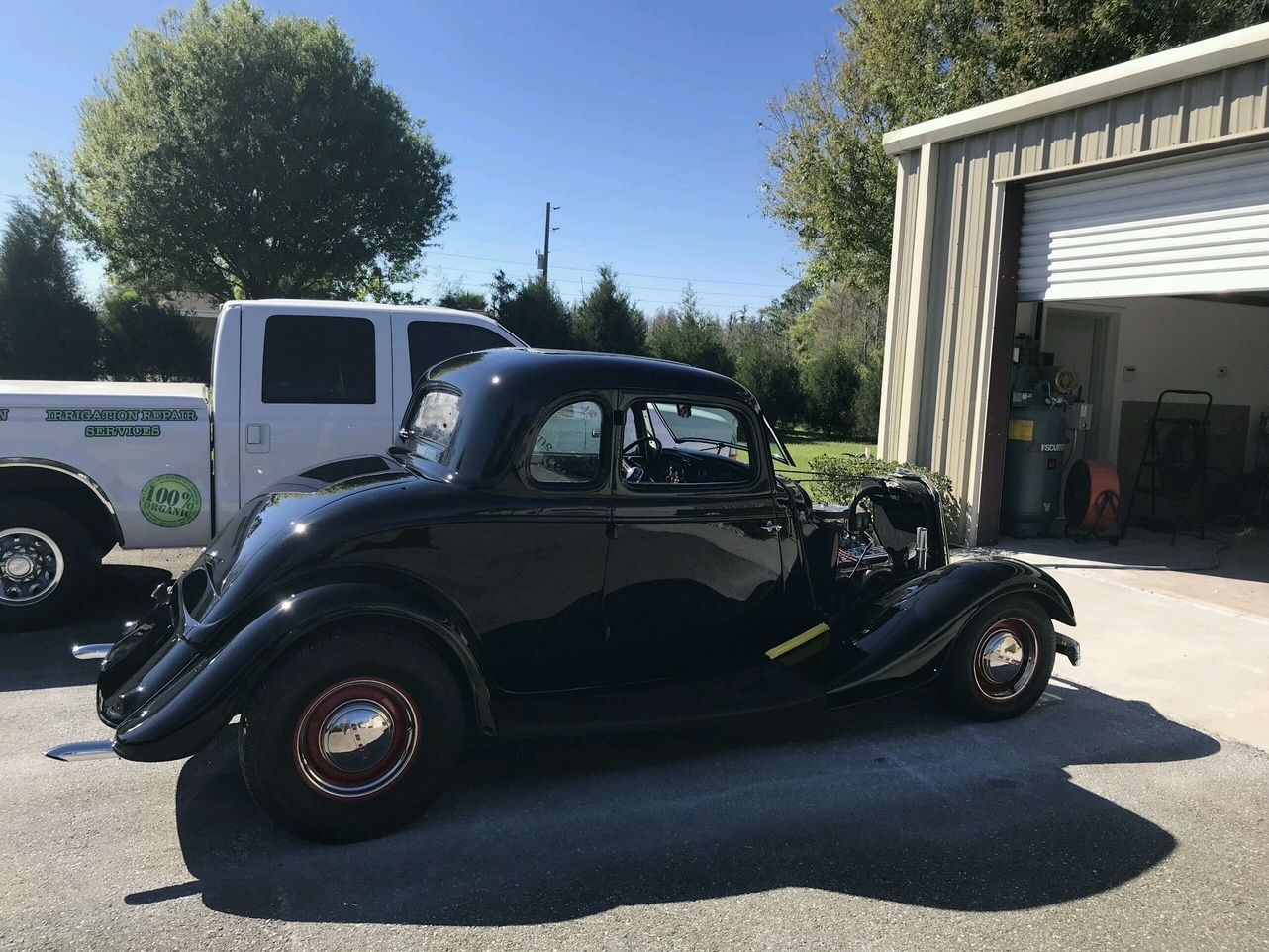 A black vintage 1930s Ford coupe parked outdoors next to a white pickup truck in front of a garage.