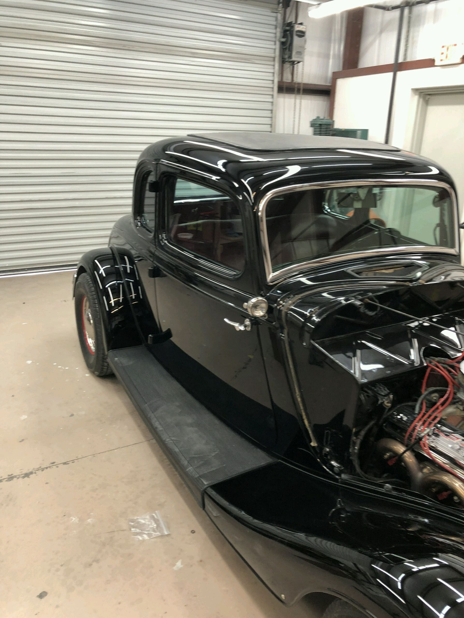 A black vintage hot rod coupe with chrome trim, parked inside a garage with the hood removed to reveal the engine.