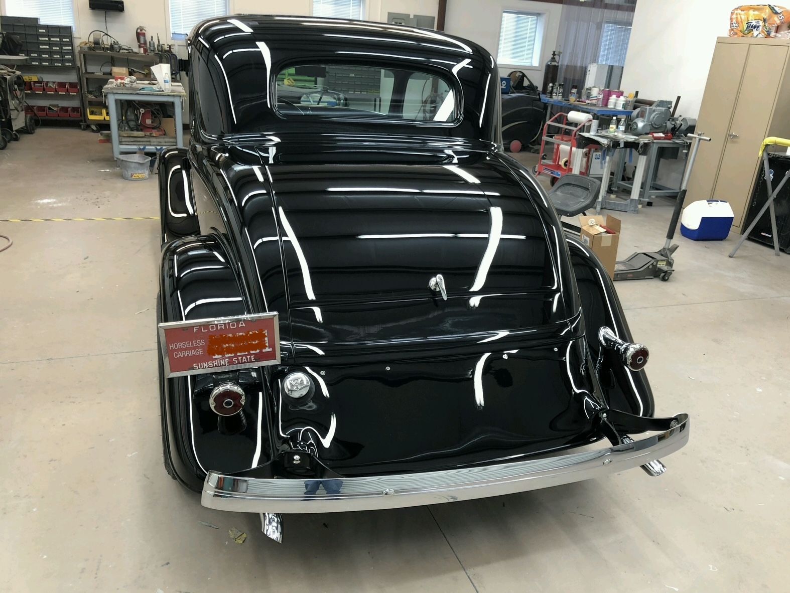 A shiny black vintage car parked in a workshop, featuring a chrome bumper and a red license plate on the rear.