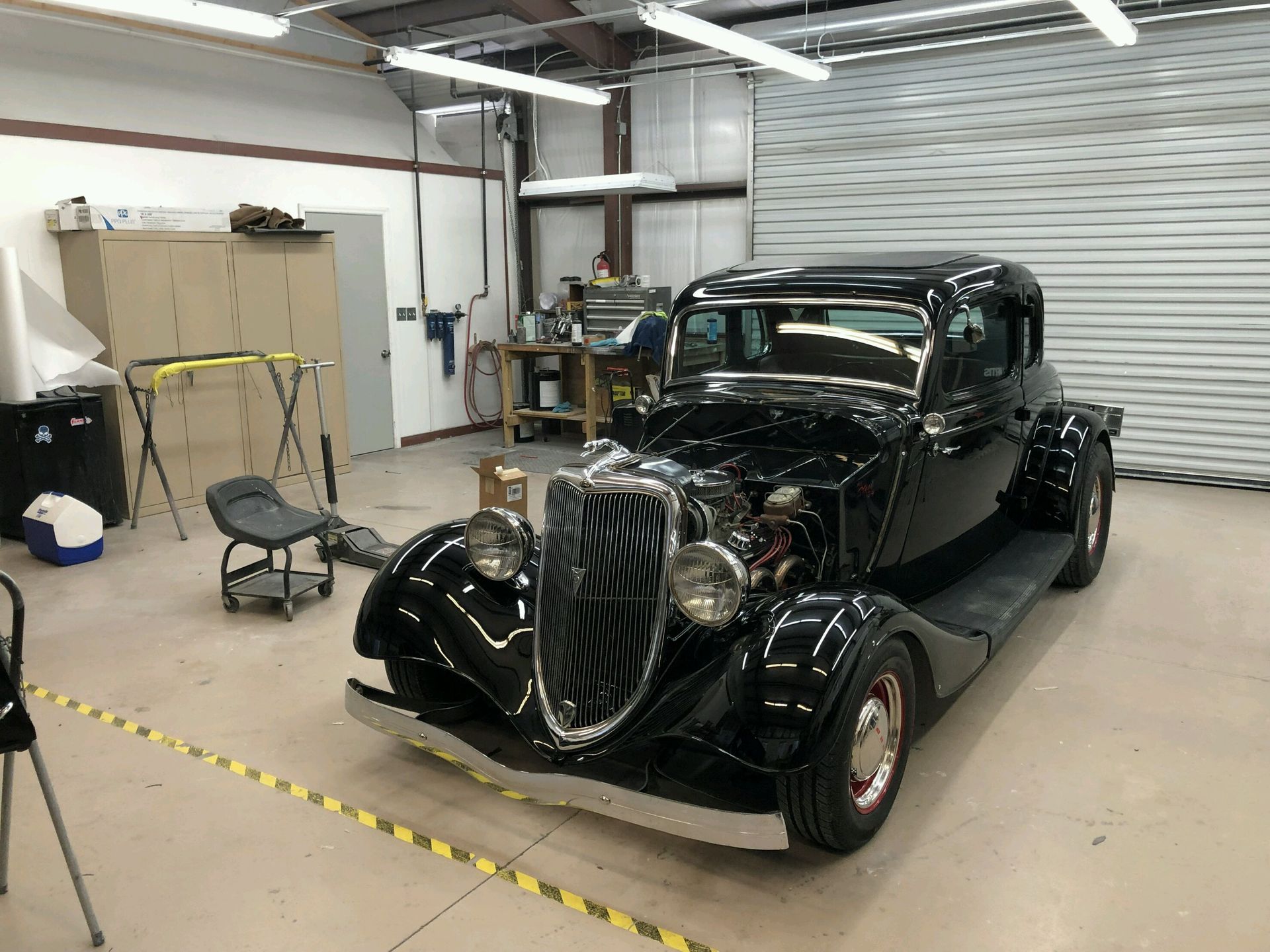 A black vintage car sits in a well-lit workshop with tools, a rolling seat, and a large metal garage door in the background.