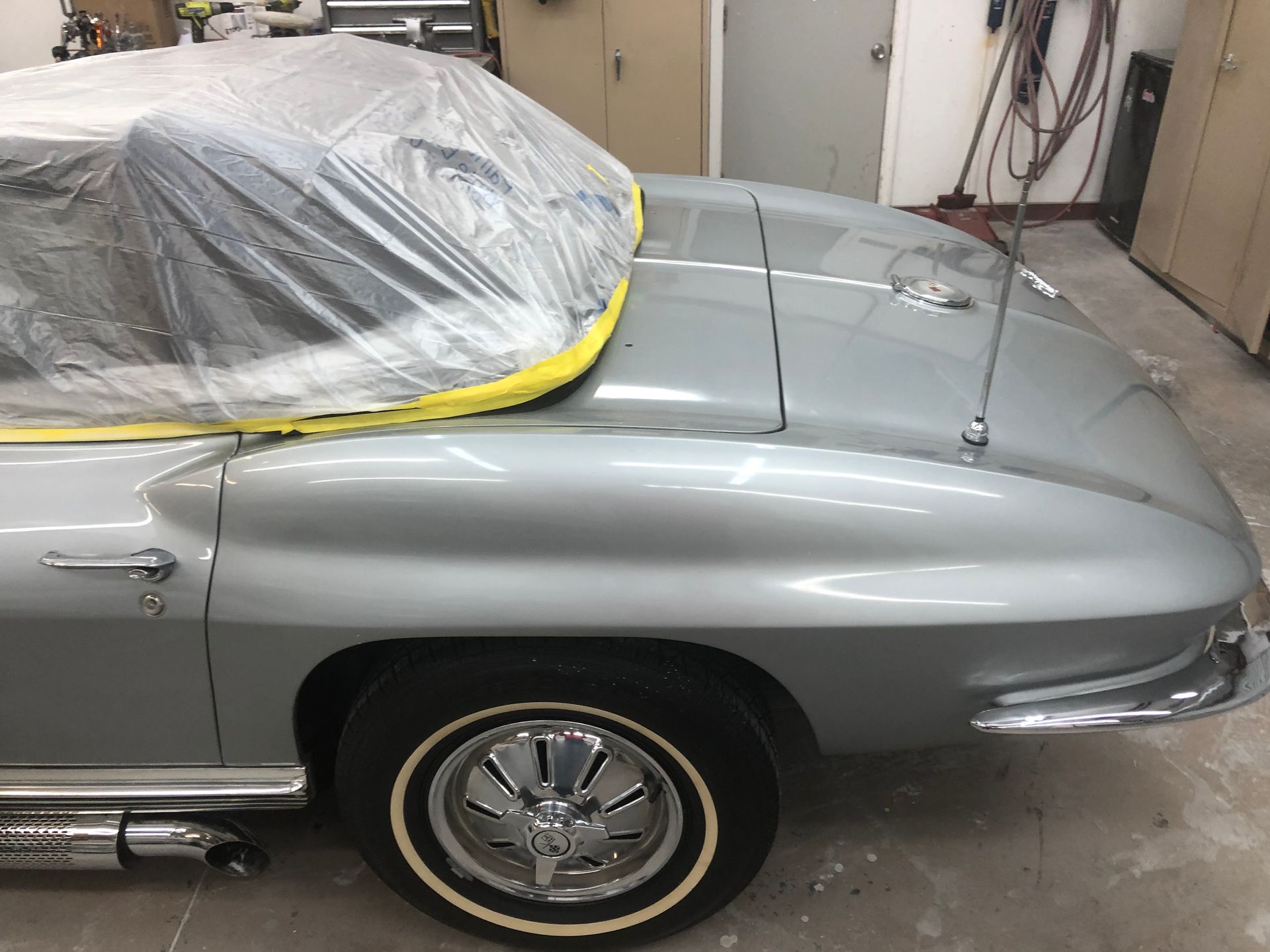 A silver classic Corvette in a garage, with a plastic cover over the convertible top and side exhaust pipes.