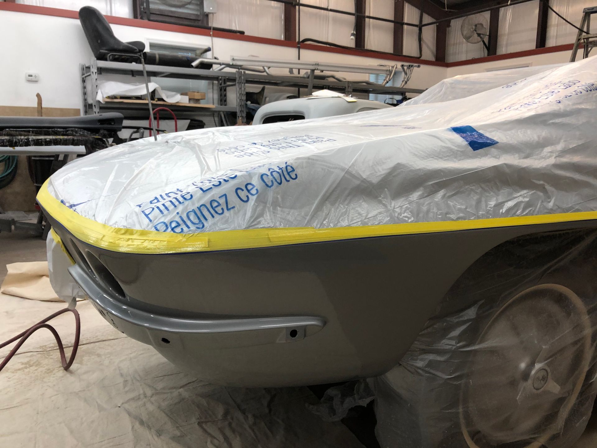 A vintage car partially covered in protective plastic and yellow masking tape in an auto body workshop.