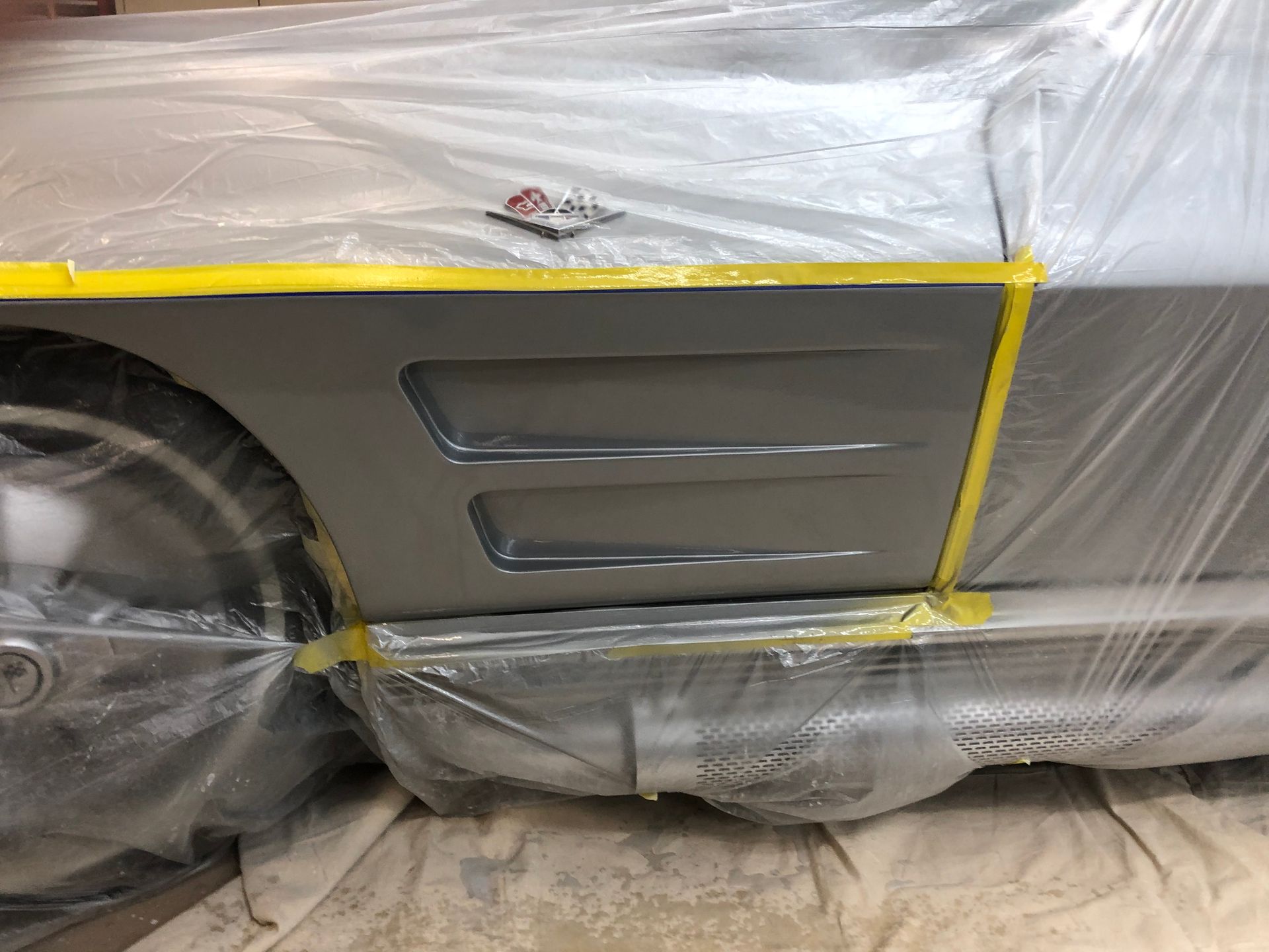 Silver car side panel with vent, covered in plastic and marked with yellow painter's tape for painting.